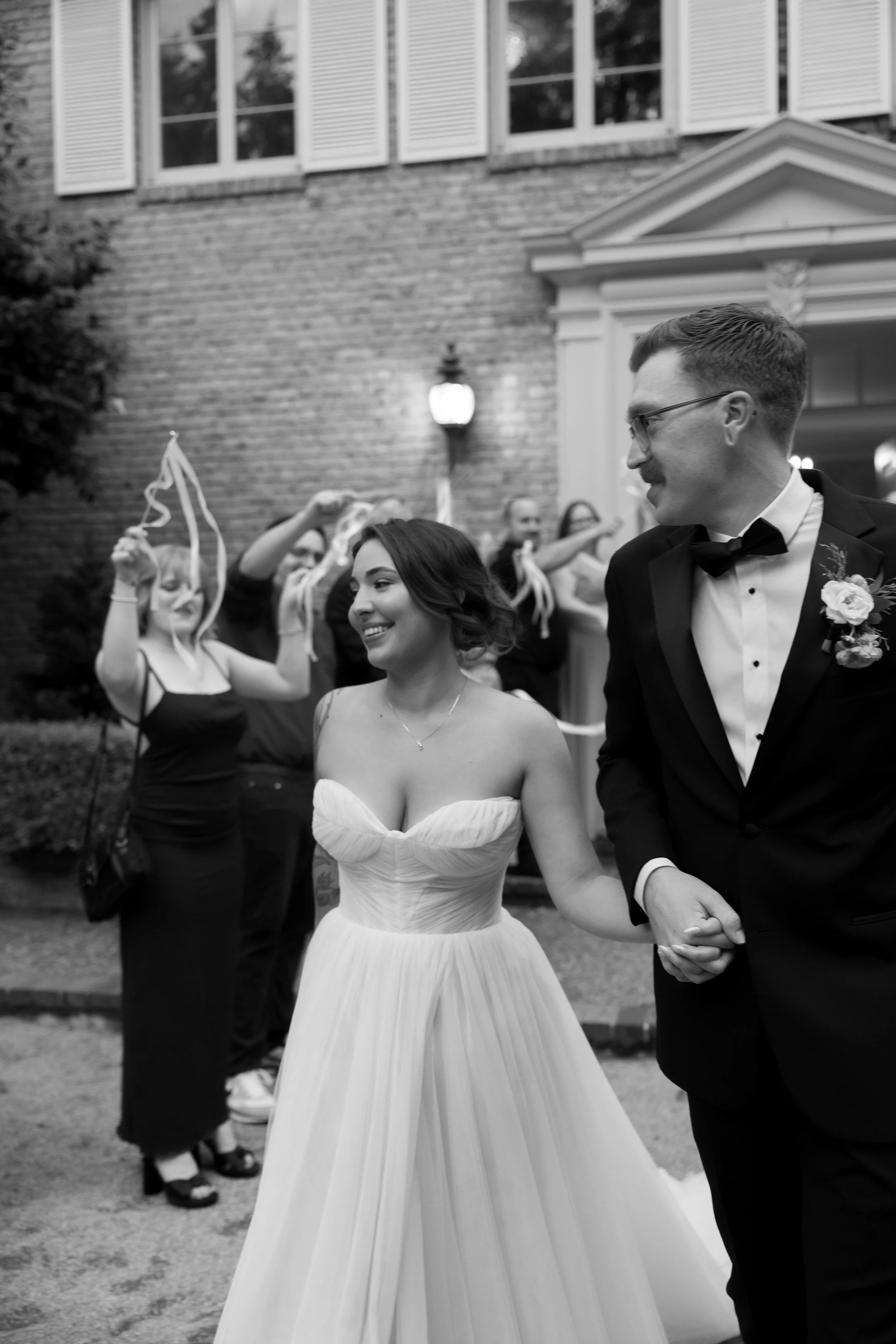 A black-and-white photo of a bride and groom holding hands and smiling, with guests throwing ribbon wands in the background at a wedding outside at a European inspired Lakewold Gardens wedding in Seattle