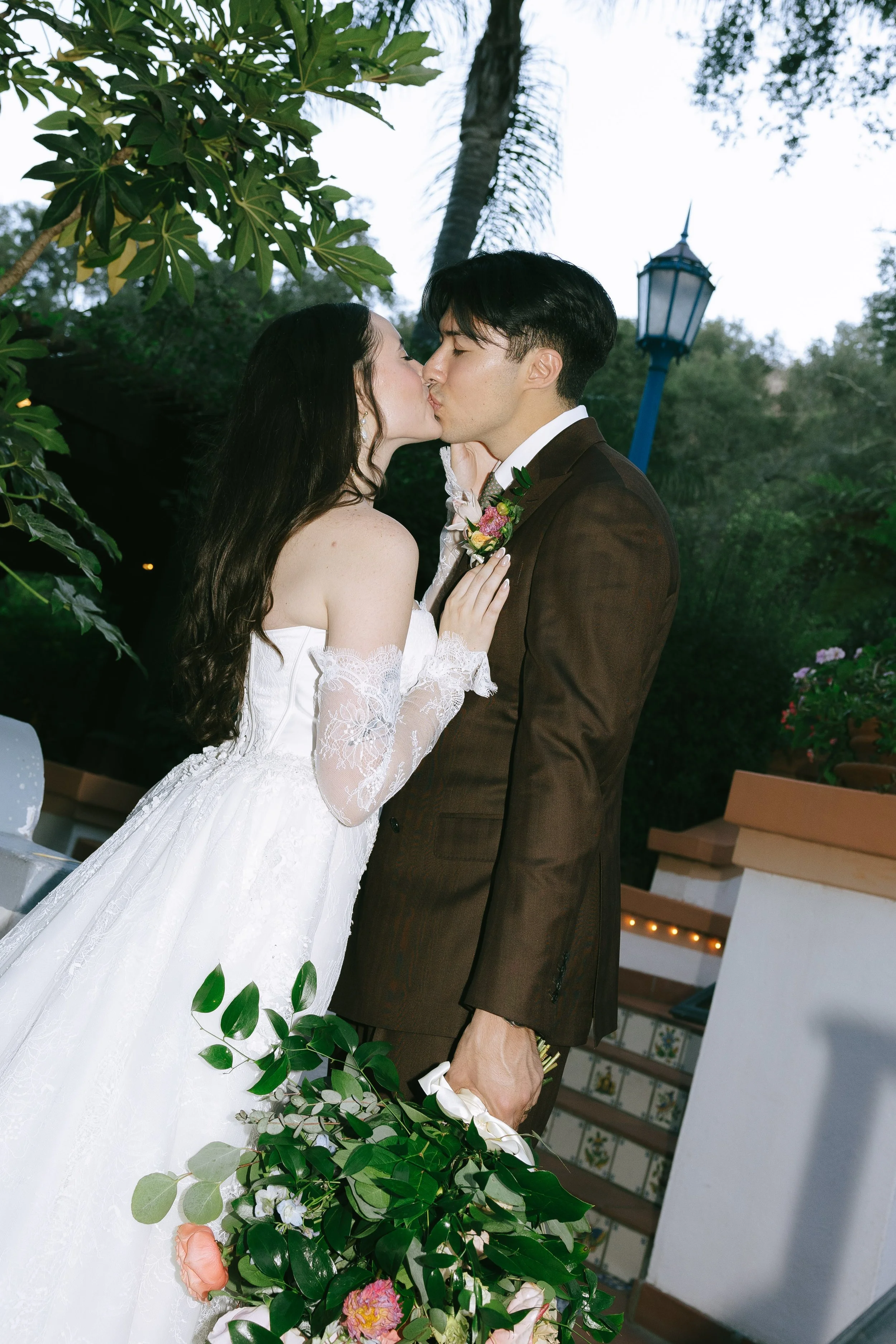 A bride and groom kissing outdoors during their wedding ceremony, with greenery and a lamp post in the background.