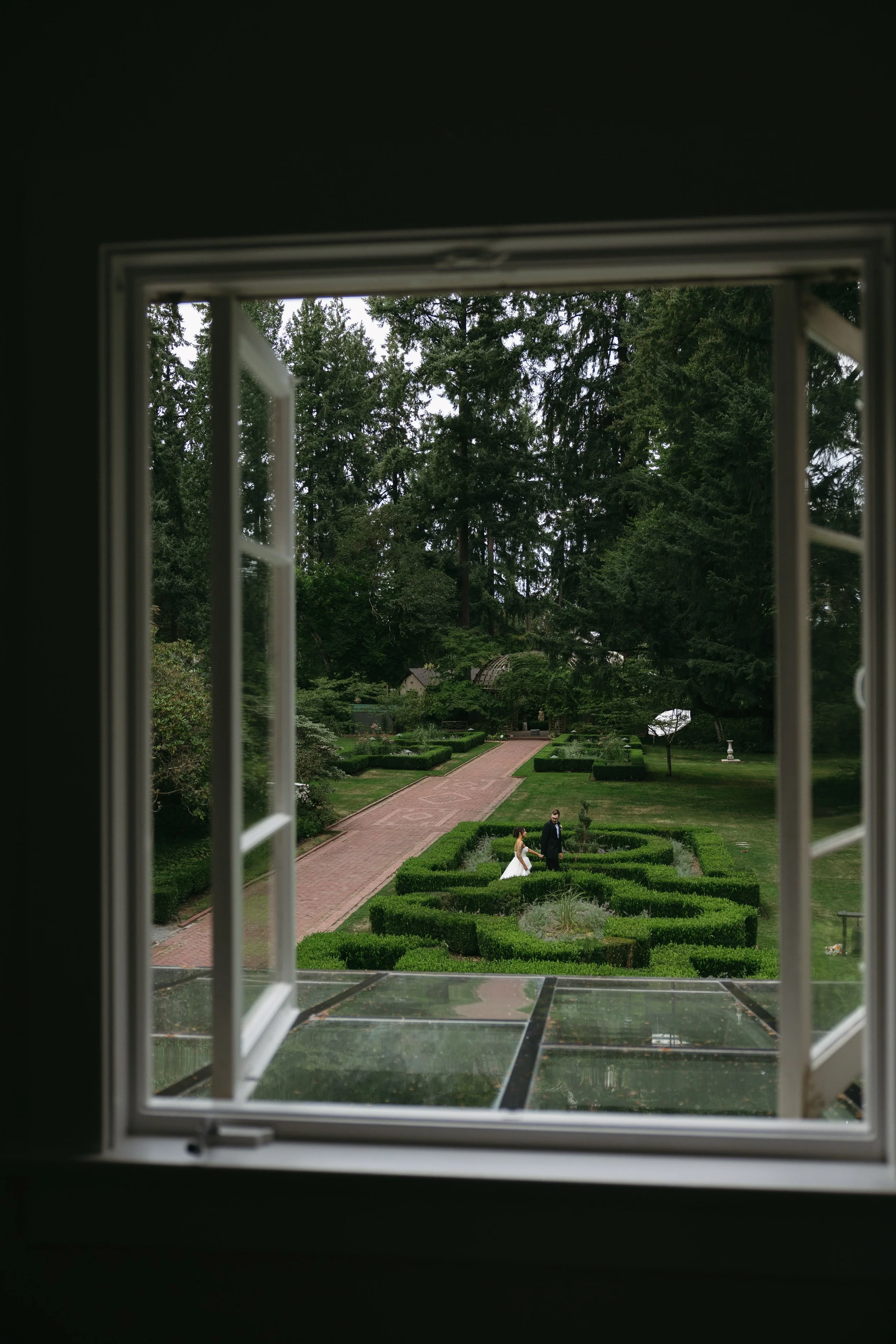 View of a wedding couple walking in a garden as seen through an open window at an European inspired wedding at Lakewold Gardens in Seattle