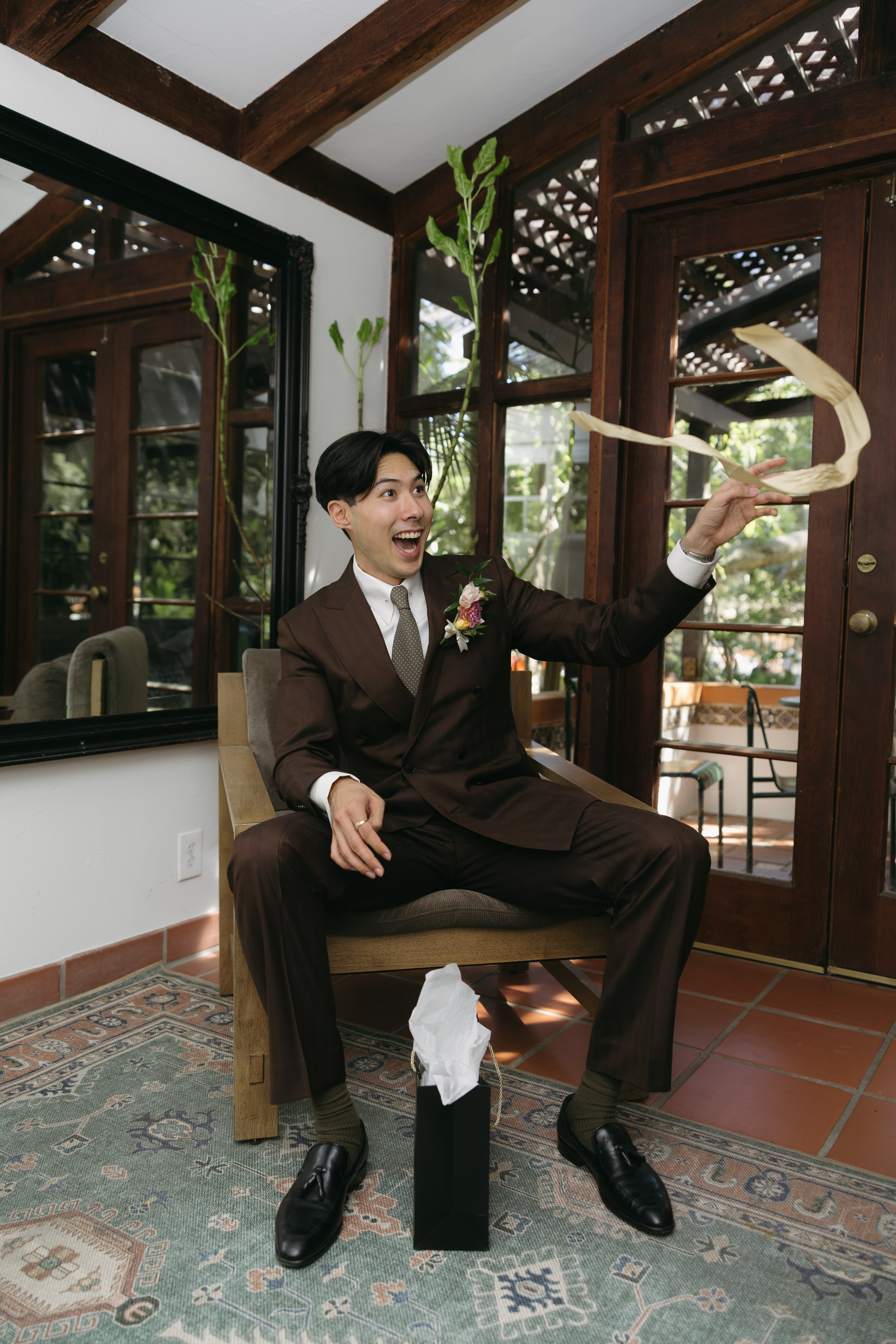 A man dressed in a brown suit with a floral boutonniere is sitting on a beige armchair inside a room with wooden framing and large windows. He appears to be throwing a hand towel and has a joyful expression. A box of tissues is on the floor in front 