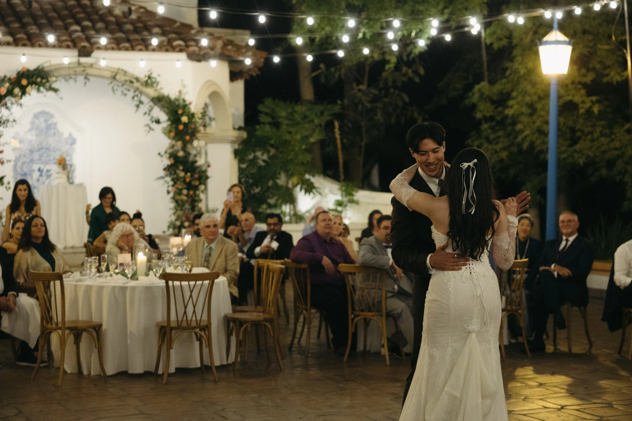 Couple dancing at a wedding reception outdoors at night, with guests seated at round tables watching, string lights overhead, and a decorative floral arch in the background.