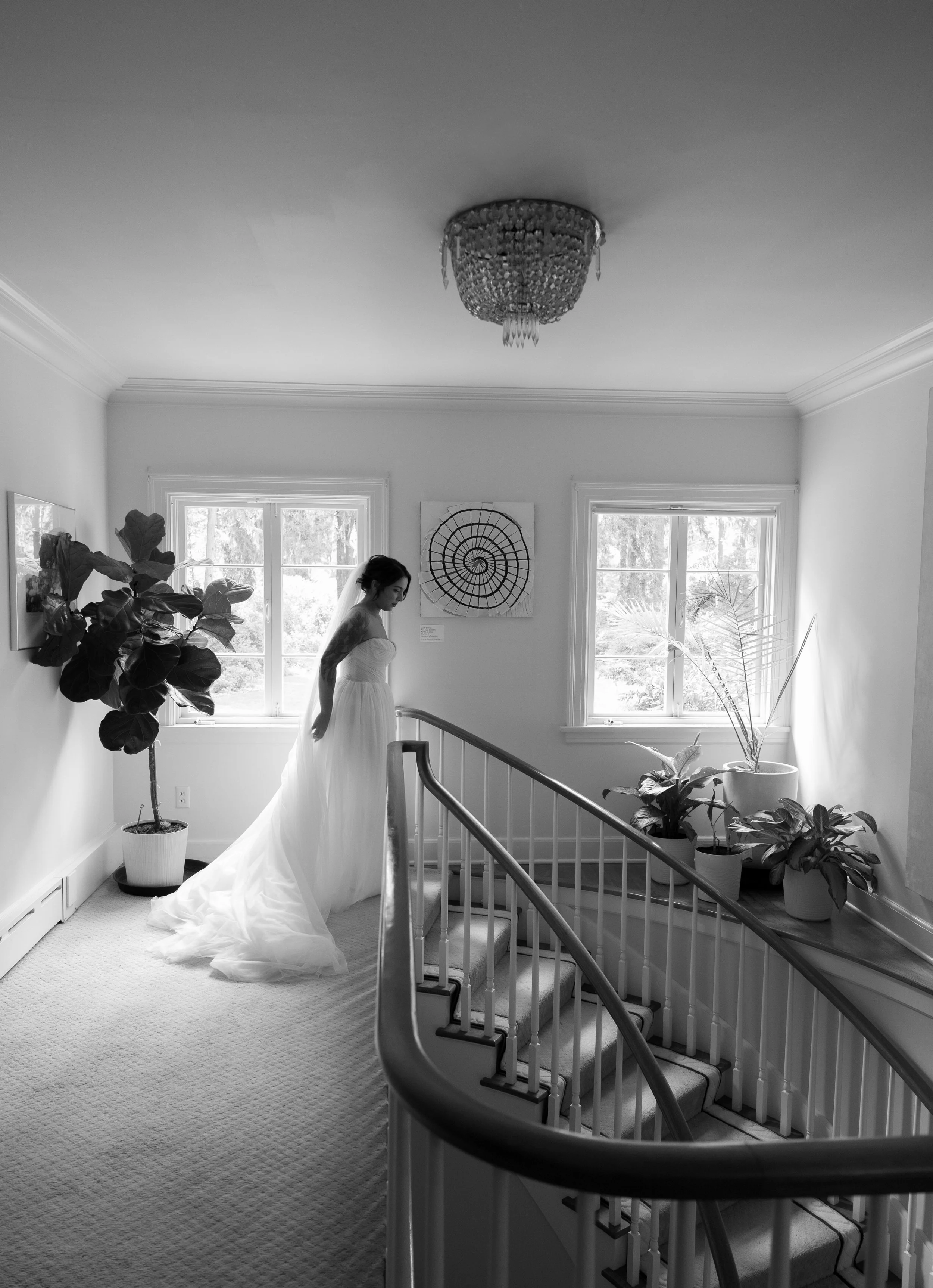 A bride in a wedding dress standing near the top of a staircase in a bright, well-lit room with large windows and potted plants at a European inspired Lakewold Gardens wedding in Seattle