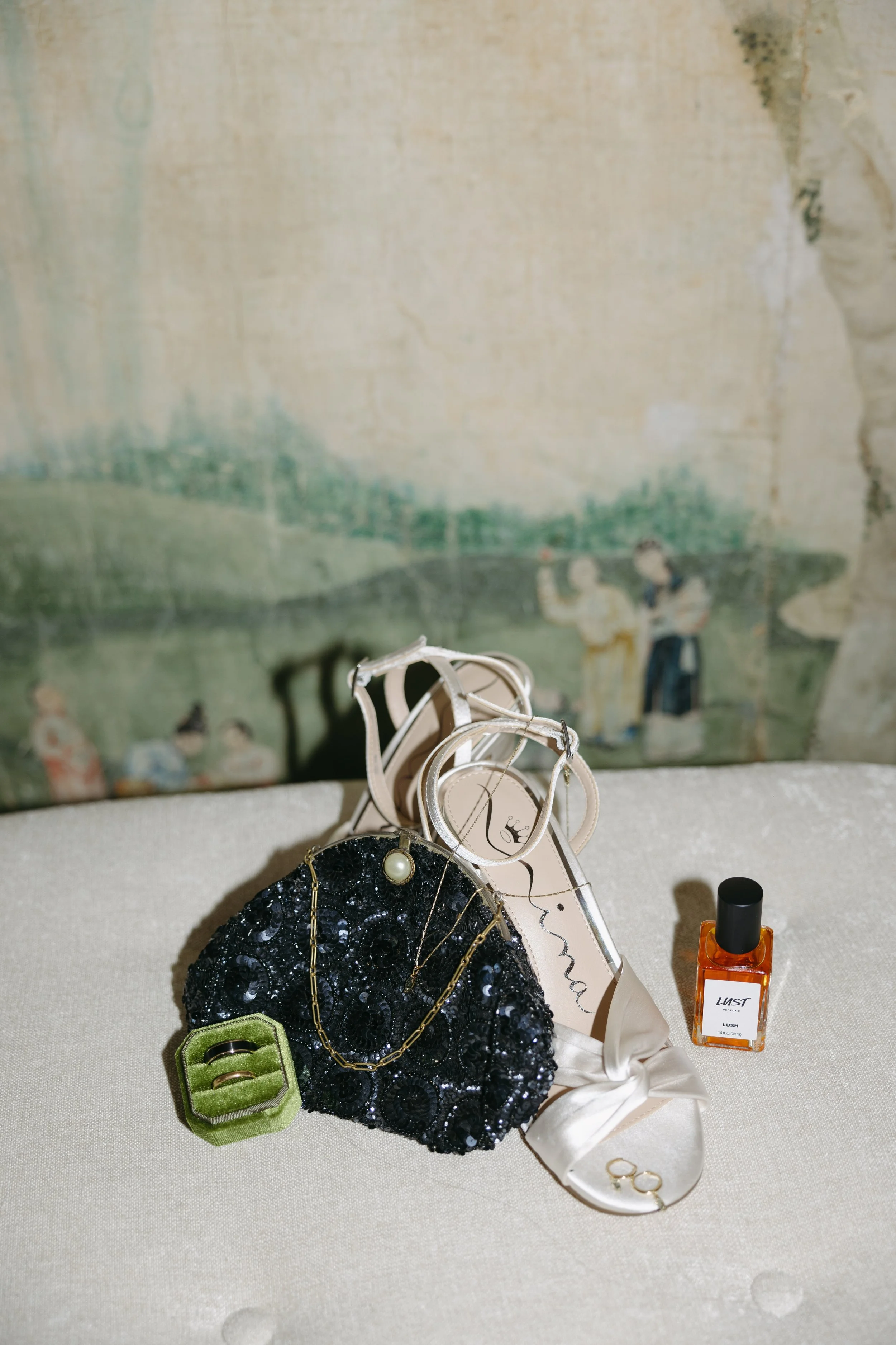 Assorted jewelry and accessories including a black sequined clutch, a gold chain necklace with a pearl, two rings in a green velvet holder, a pair of white satin shoes, a small perfume bottle labeled 'Lush,' and a pair of sunglasses, placed on a beig