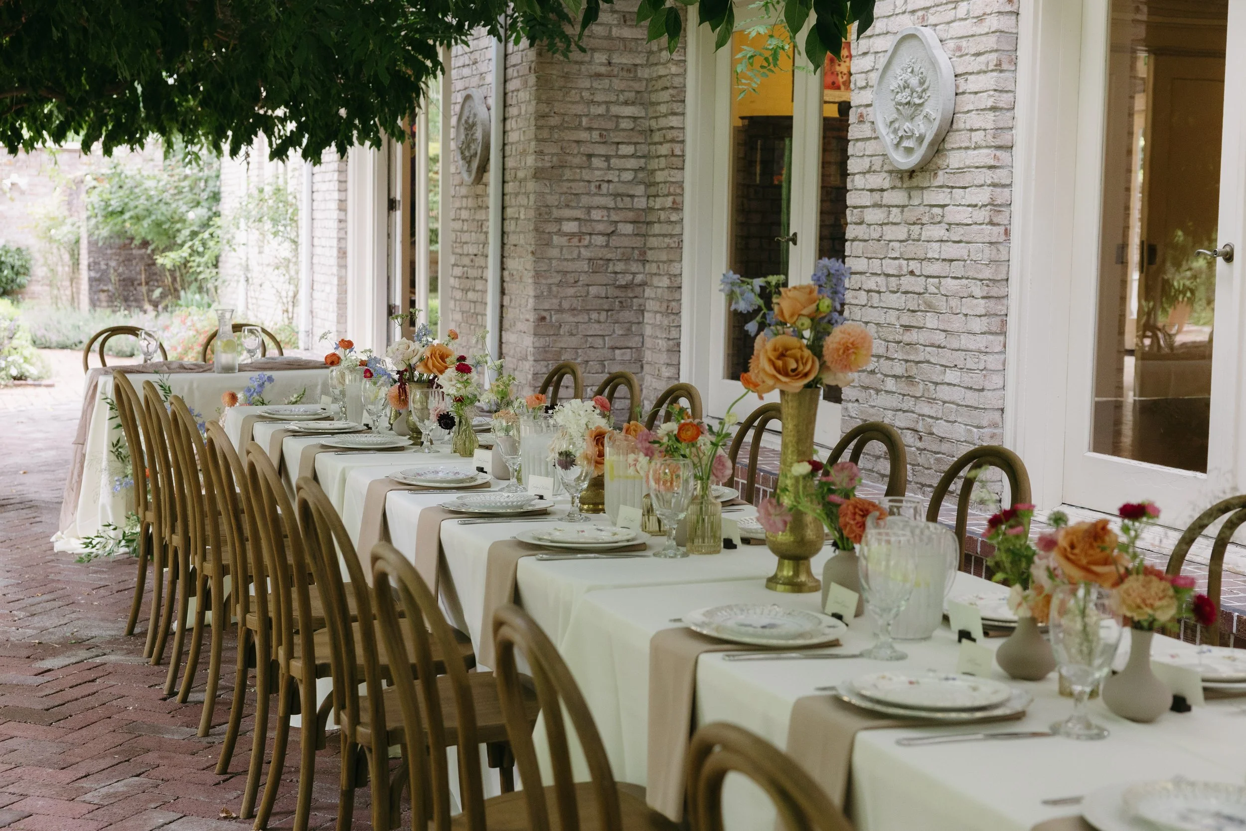 Long outdoor dining table set with white tablecloths, floral centerpieces, plates, glassware, and napkins, adjacent to a brick wall with windows and decorative wall plaques, under trees with green leaves.