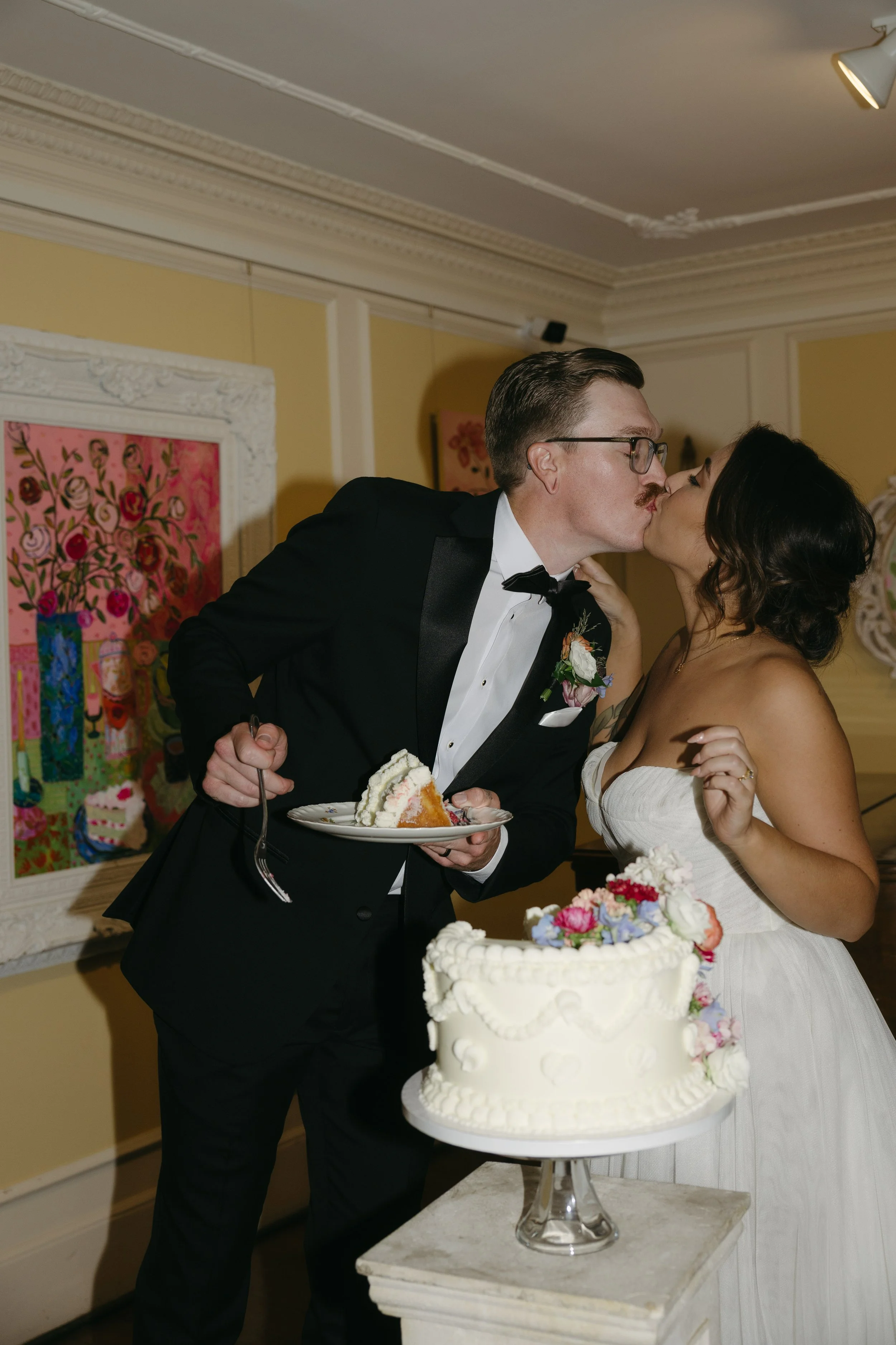 A newlywed couple sharing a kiss during their wedding celebration, with a wedding cake on a table in front of them.