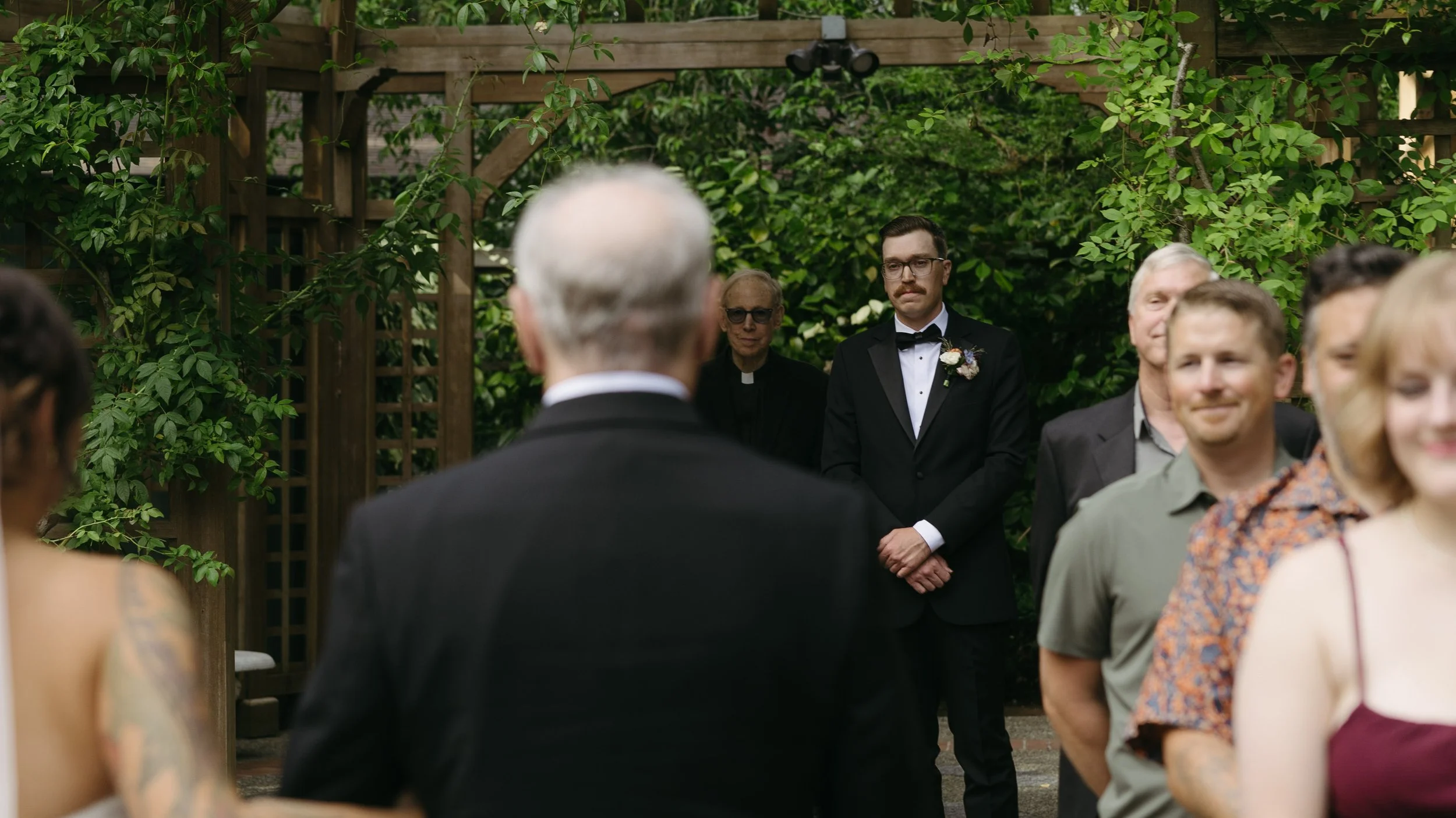 A groom in a black tuxedo standing with a floral boutonniere, facing a man with gray hair dressed in black. They are in an outdoor garden setting with lush greenery and wooden structures in the background, while guests look on.