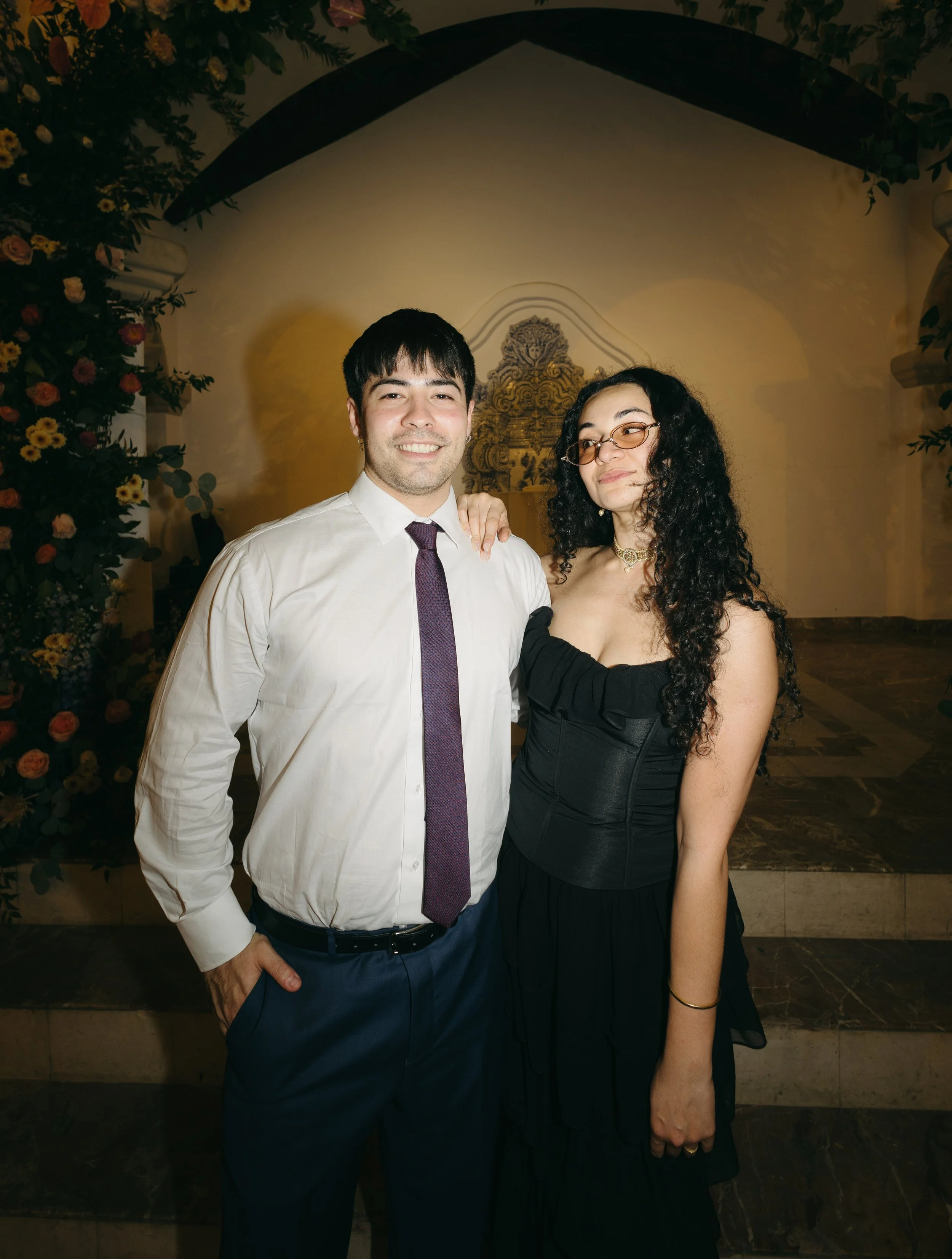 Young man and woman standing together indoors, dressed stylishly, with floral decorations in the background.