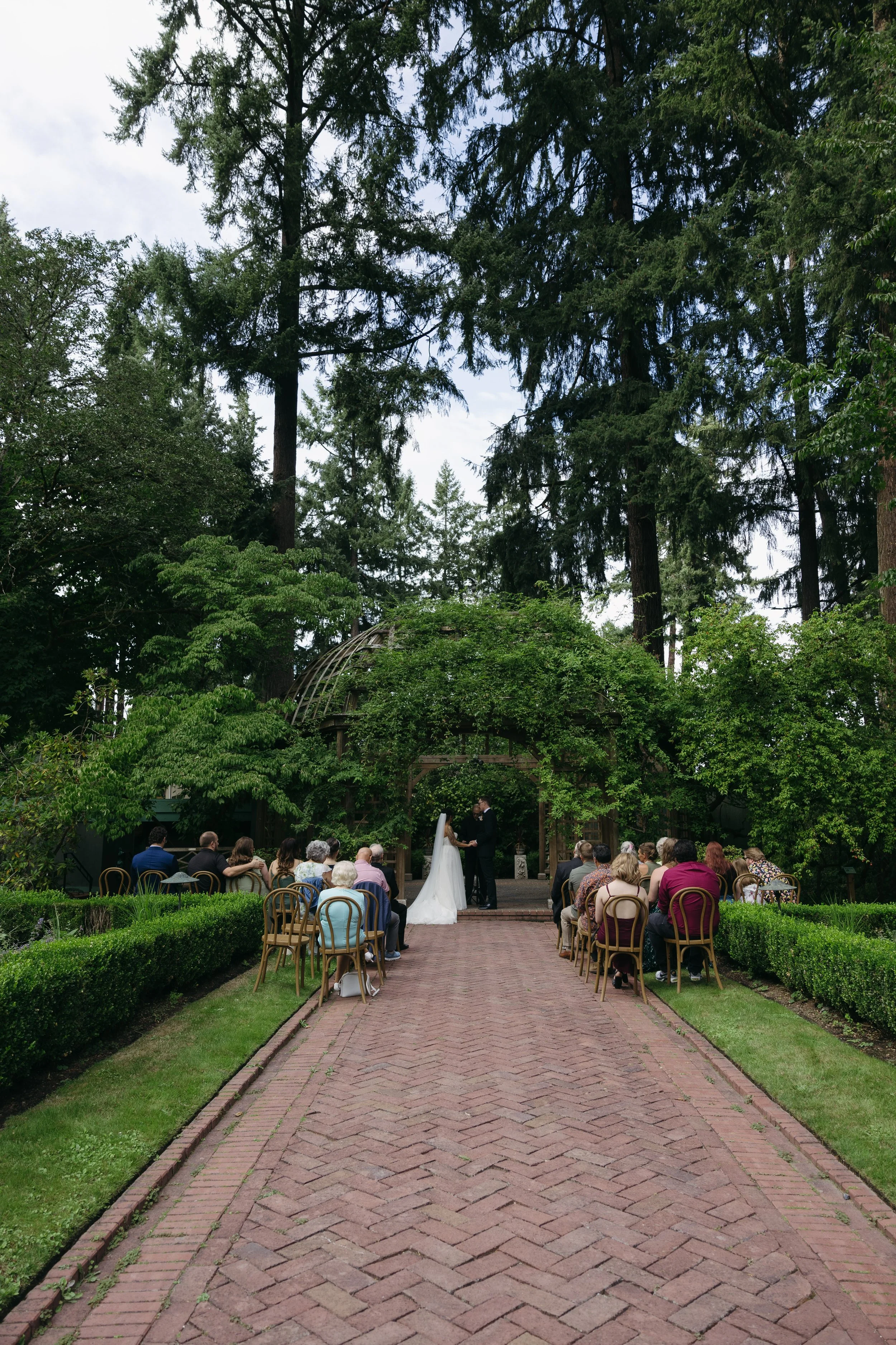 A wedding ceremony taking place outdoors on a brick pathway, surrounded by greenery and tall trees, with guests seated on chairs and the bride and groom standing at the altar at a European inspired Lakewold Gardens wedding in Seattle