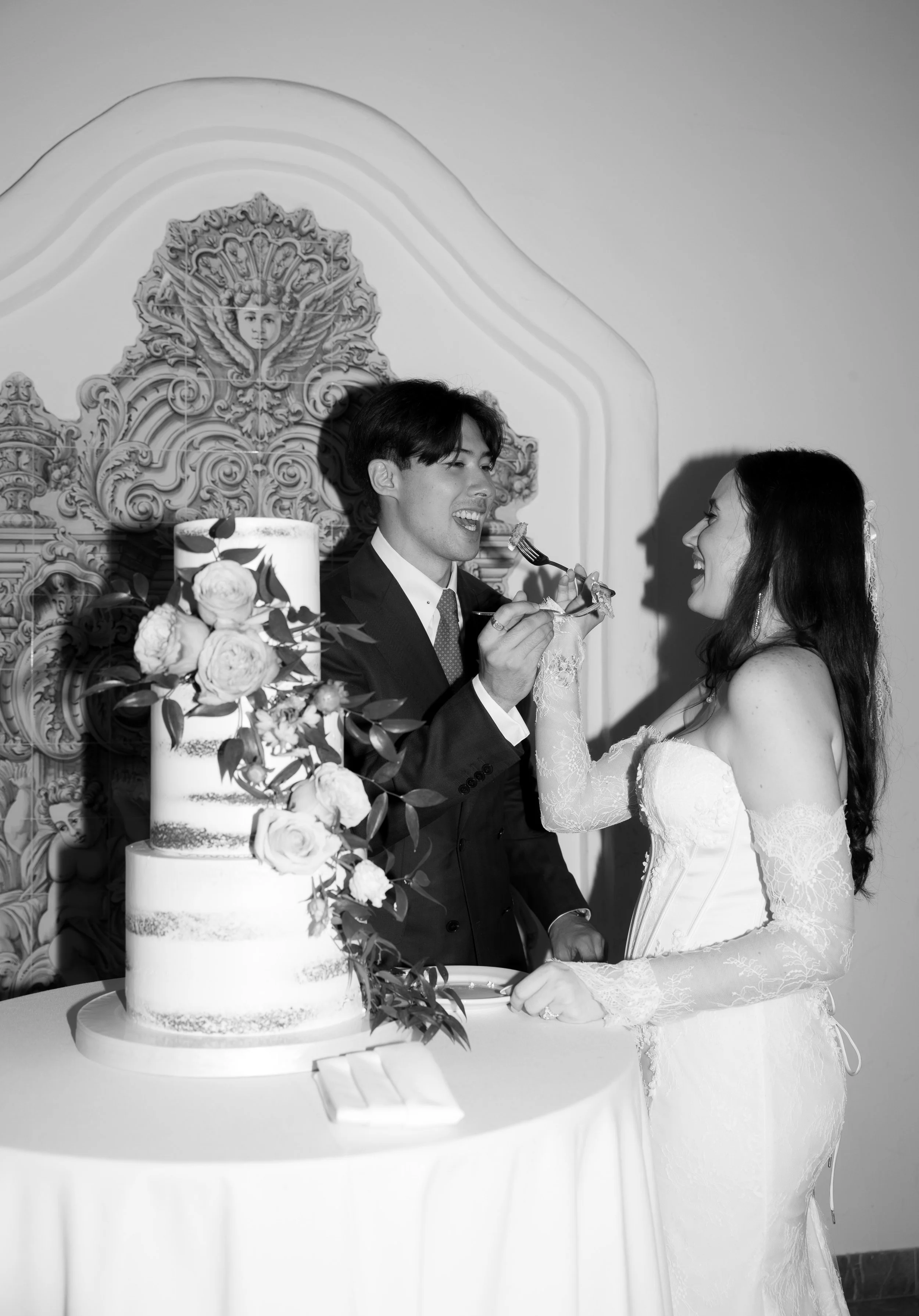 A bride and groom at their wedding cake, sharing a moment of feeding each other, smiling, with a decorated cake and ornate backdrop in the background.