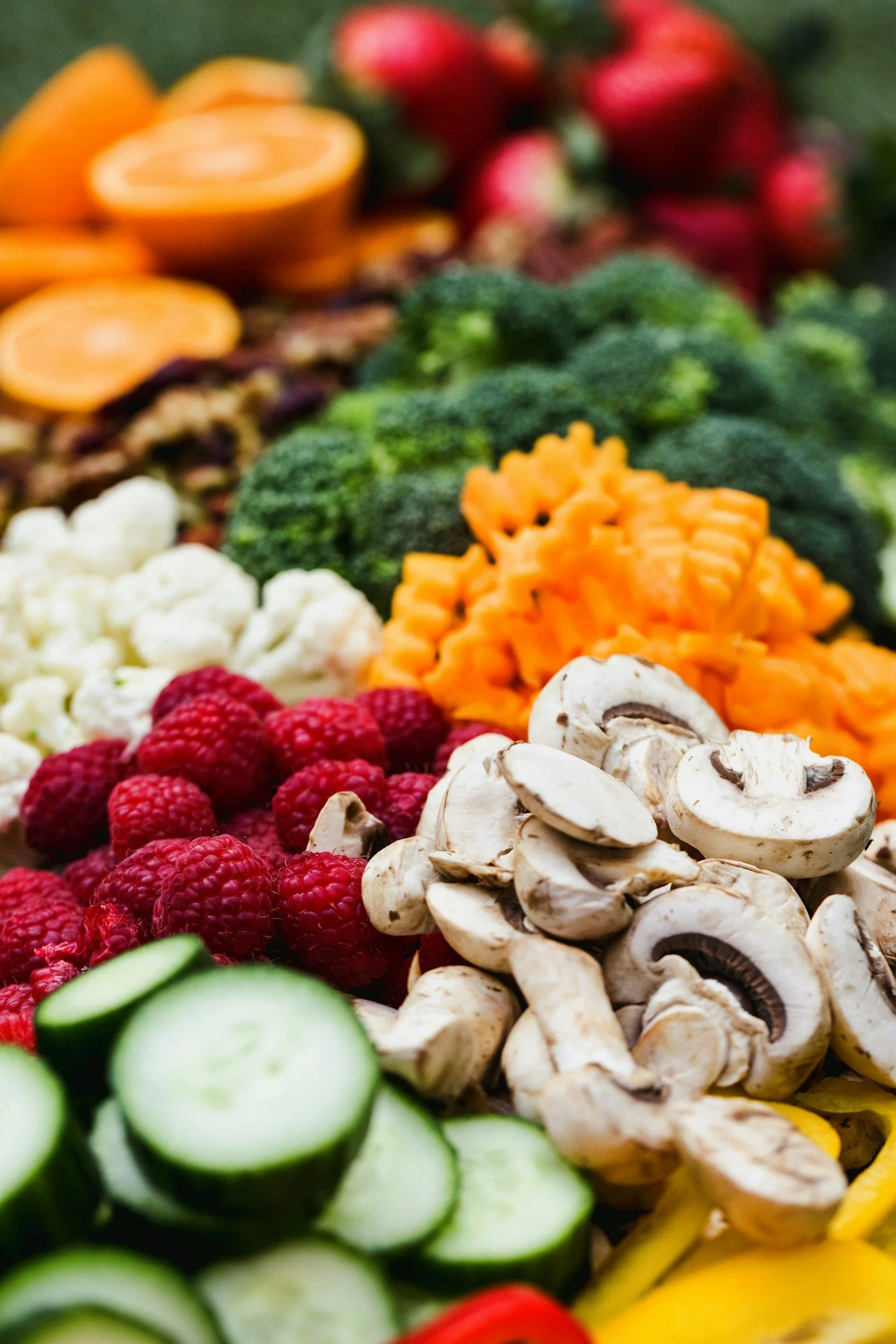 Assorted fresh vegetables and fruits including sliced cucumbers, raspberries, sliced mushrooms, shredded carrots, broccoli florets, sliced strawberries, and orange slices.