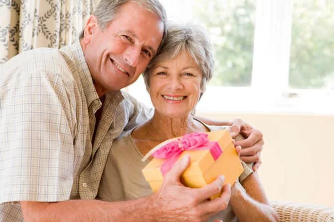 An elderly couple smiling and embracing while holding a gift with a pink ribbon.