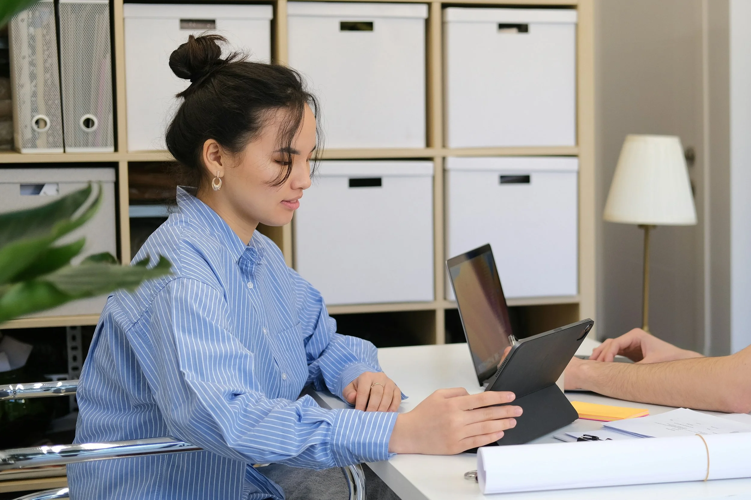 A woman in a blue striped shirt sitting at a desk, looking at her tablet in a case, in an office with shelves, a lamp, and plants.