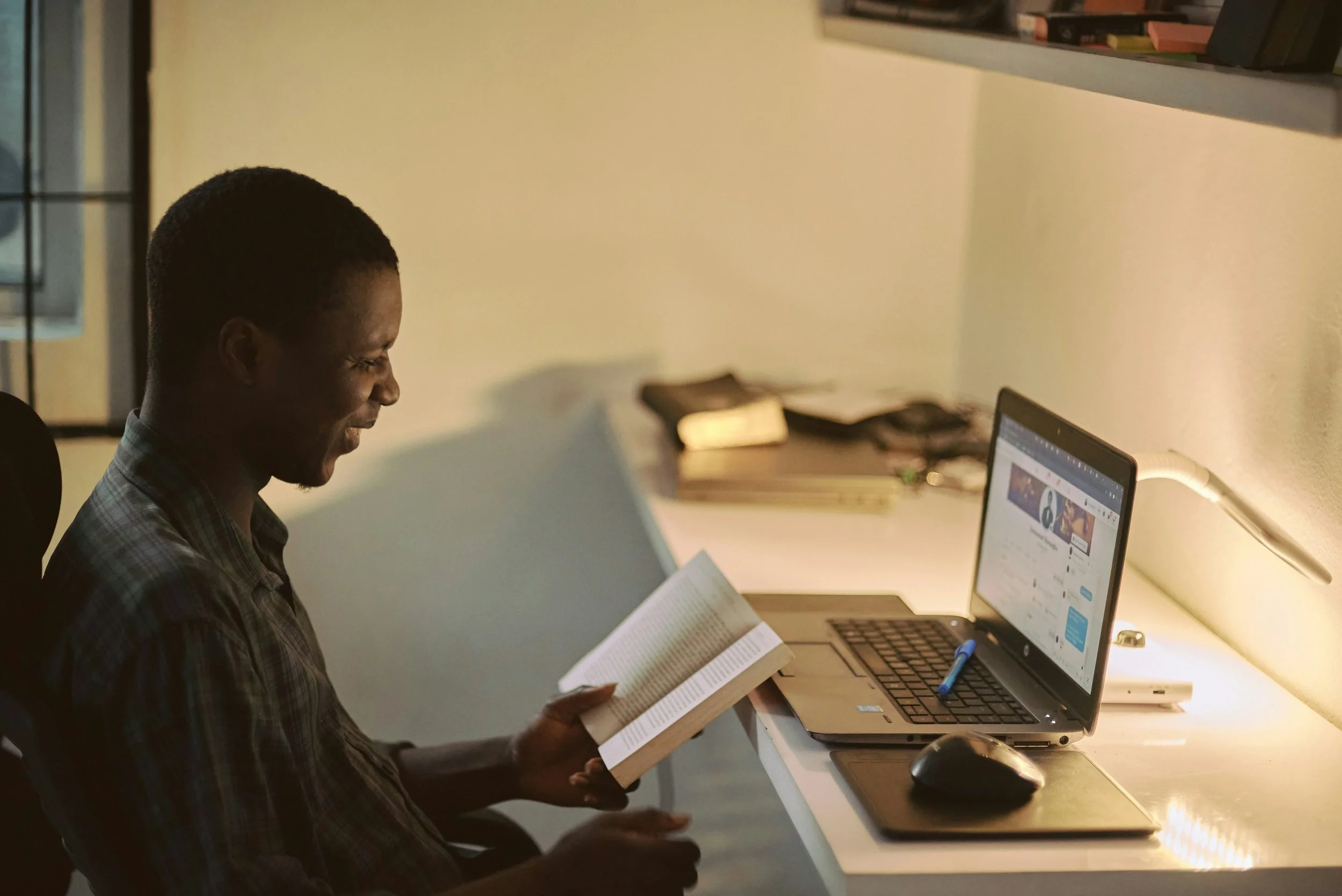 A young man sitting at a desk, reading a book, with a laptop, mouse, and a lamp on the desk, in a well-lit room.