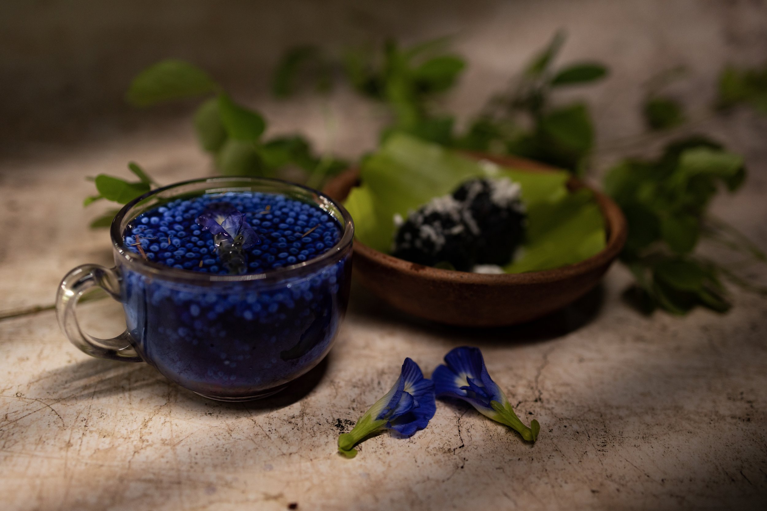 A glass cup filled with blue flower petals and tiny flower-like beads, with two blue flowers lying beside it on a rustic wooden surface. In the background, there is a brown bowl with green leaves and a piece of black and white volcanic rock.