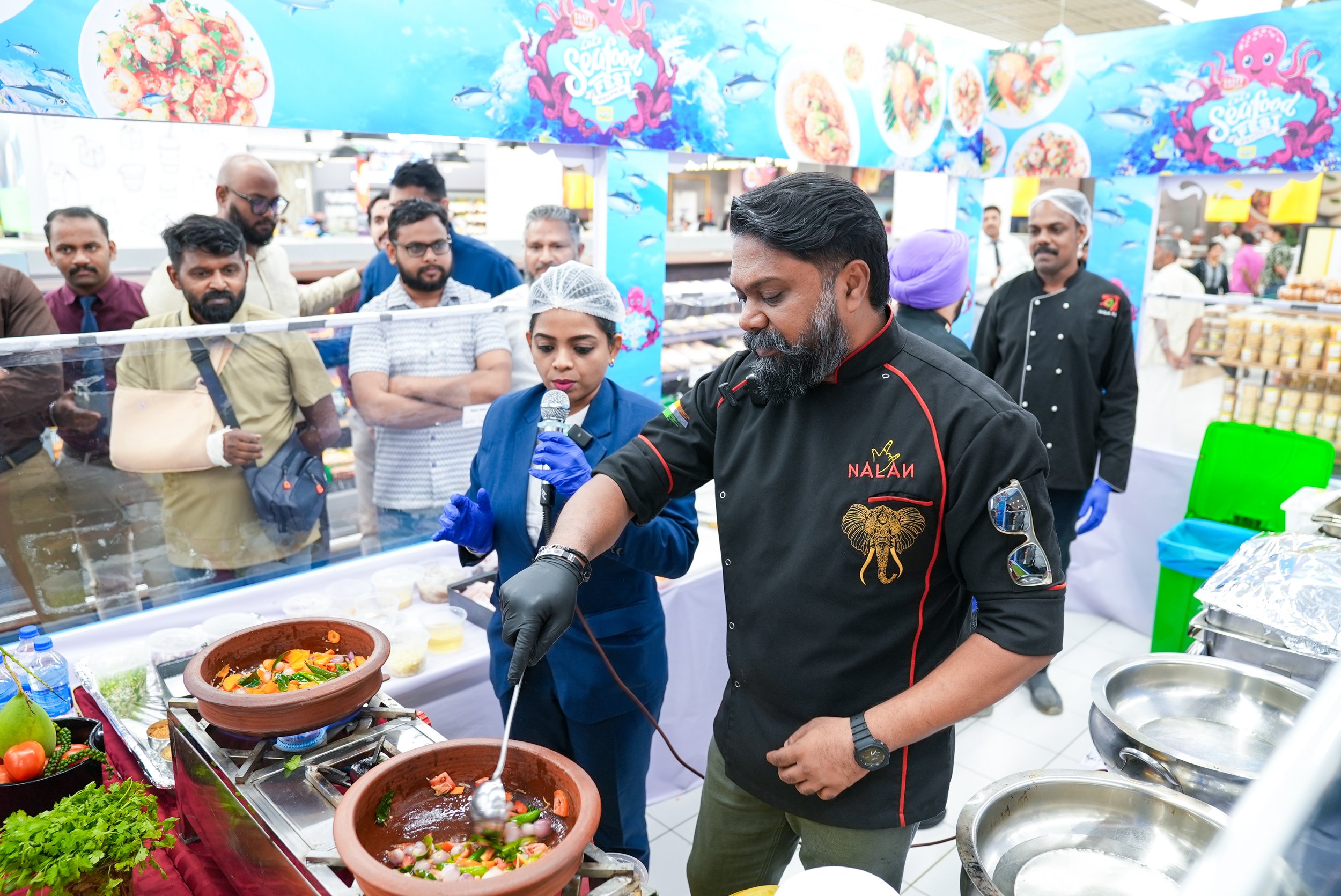 Chef demonstrates cooking seafood dishes in clay pots at a food expo while a woman with a microphone explains, and an audience observes behind a glass barrier.