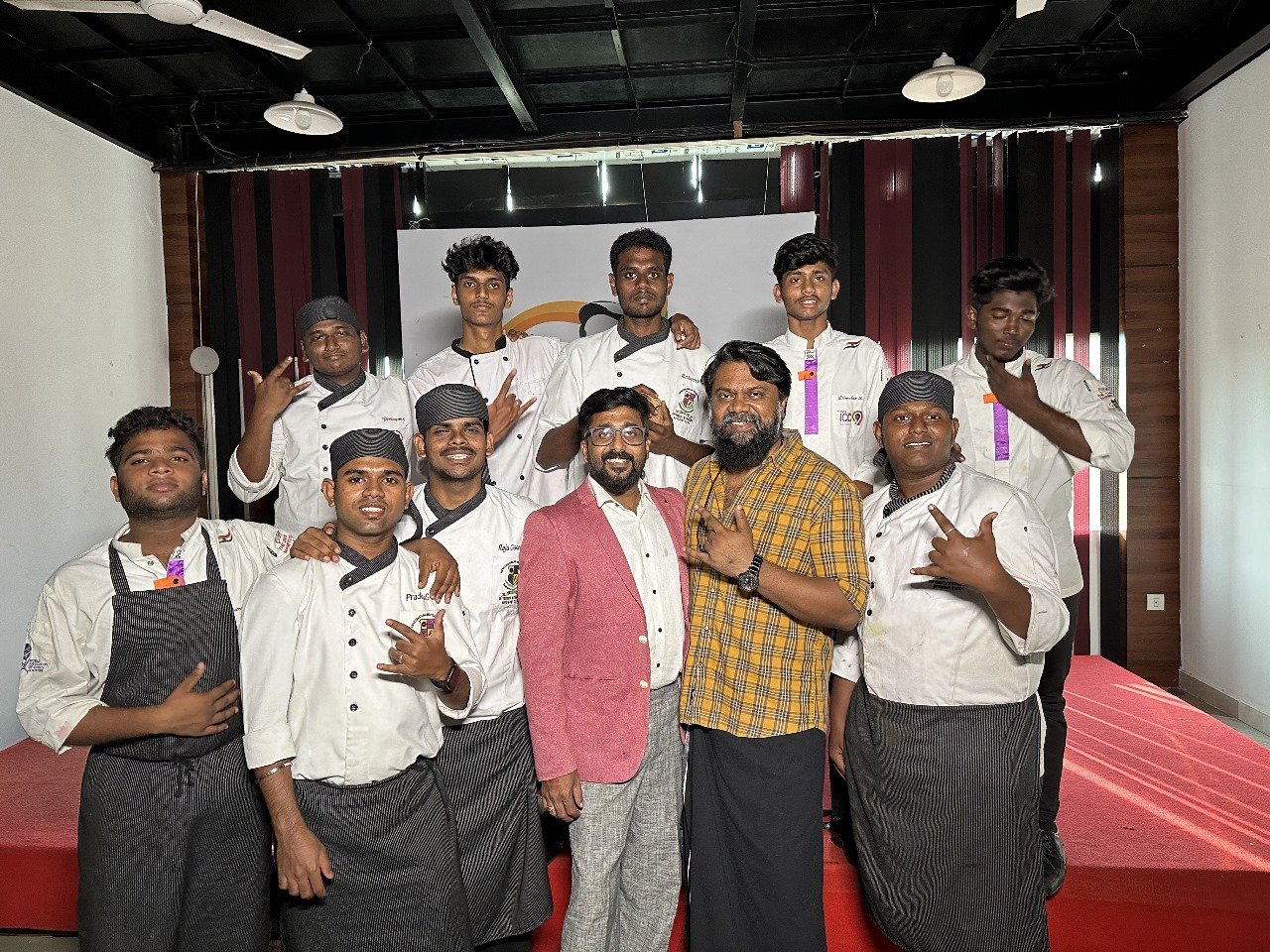 Group of eleven people, including chefs and two men in casual and formal attire, posing together on a stage with a red carpet, black and white background, and indoor lighting.