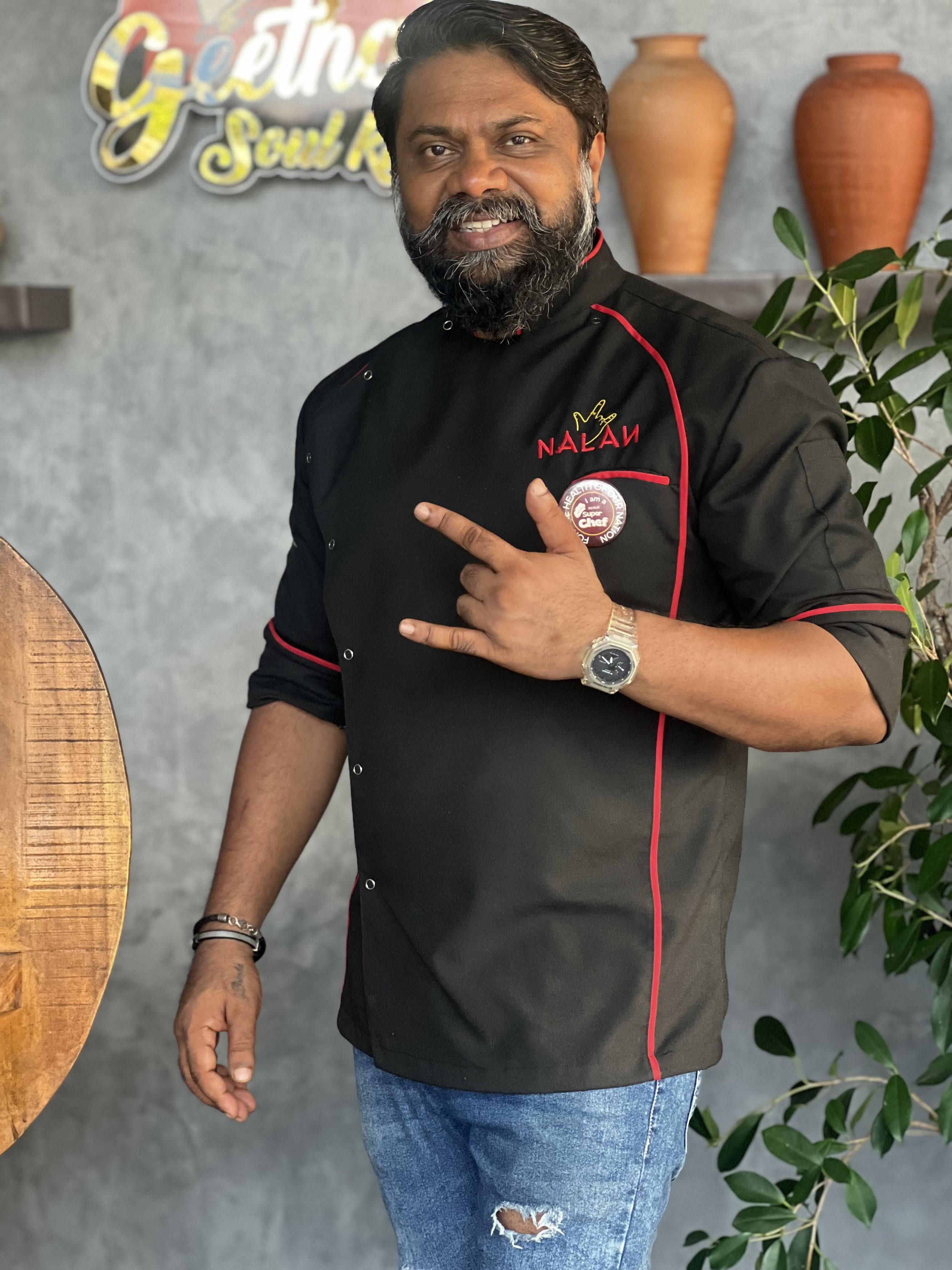 A man with a beard and mustache, wearing a black chef's coat with red piping and the name NALANI embroidered on it, standing indoors in front of a textured gray wall with decorative pottery and plants. He is pointing to his chest with his right hand, which is wearing a silver watch, and smiling.