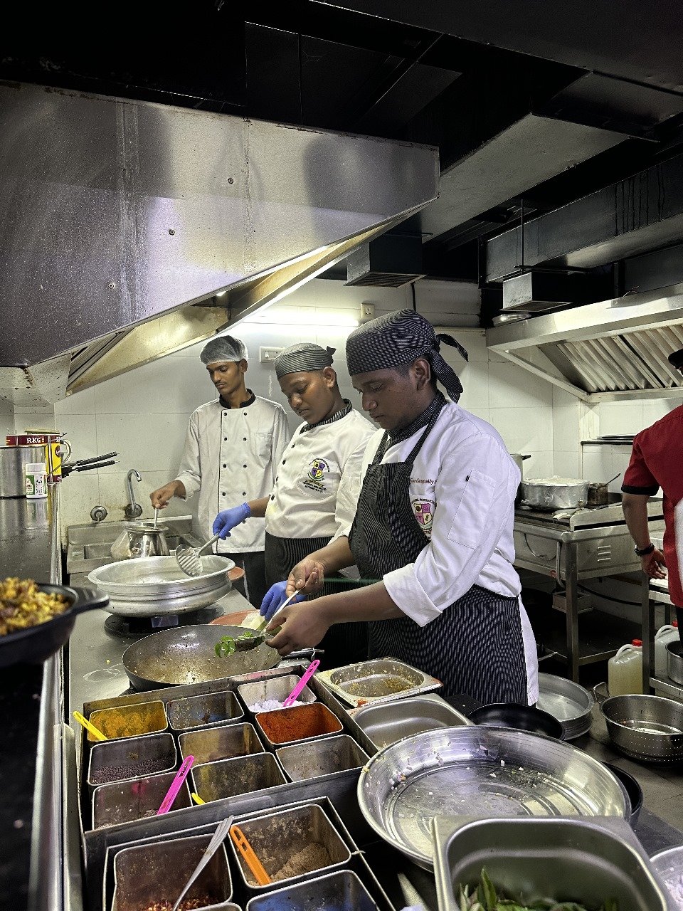 Three chefs wearing white coats and head coverings are working in a commercial kitchen, preparing and cooking food. Various containers of spices and ingredients are in the foreground.