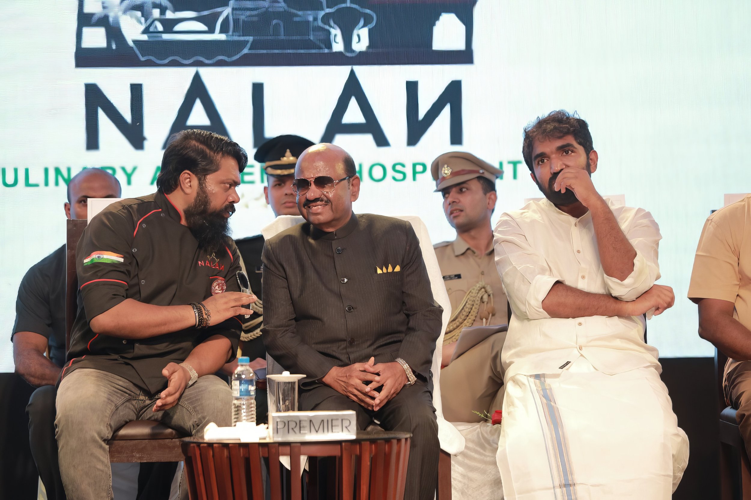 Three men sitting on stage during an event, with police officers in the background. One man on the left is wearing a black shirt with red accents, speaking and gesturing. The man in the middle is dressed in a dark suit with sunglasses, smiling, with a sign that says 'PREMIER' in front. The man on the right is wearing traditional Indian attire, with his hand near his mouth. Behind them is a screen with a logo and text.