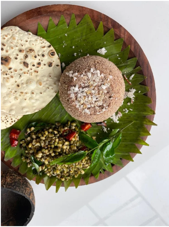 A traditional Indian meal on a round wooden plate lined with banana leaf, featuring a large papadum, a round brown bread with salt sprinkled on top, and a serving of spicy green chickpea salad with red chili slices.