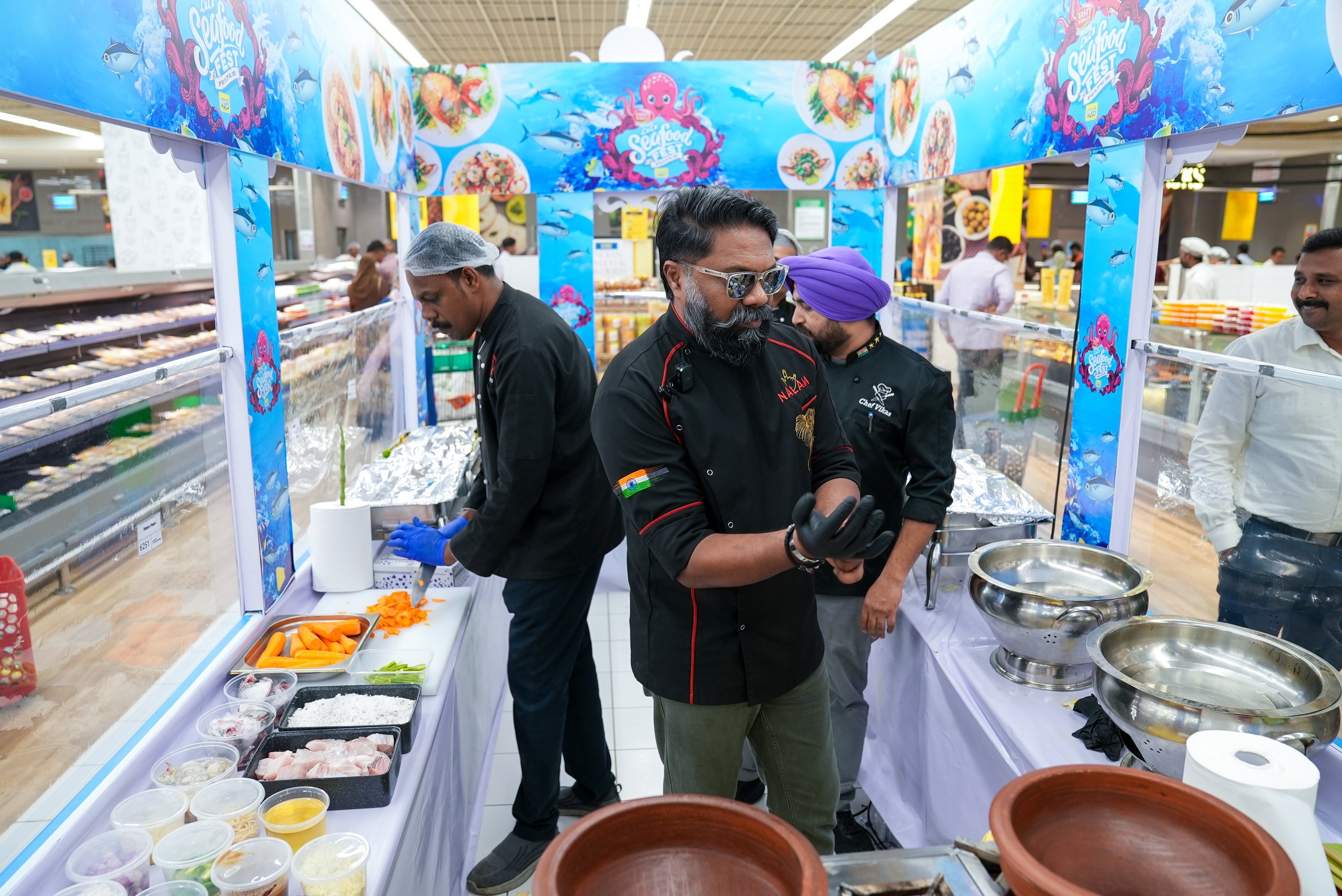 People preparing seafood dishes at a stall decorated with a blue banner featuring images of seafood, such as fish and octopus, during a food festival.