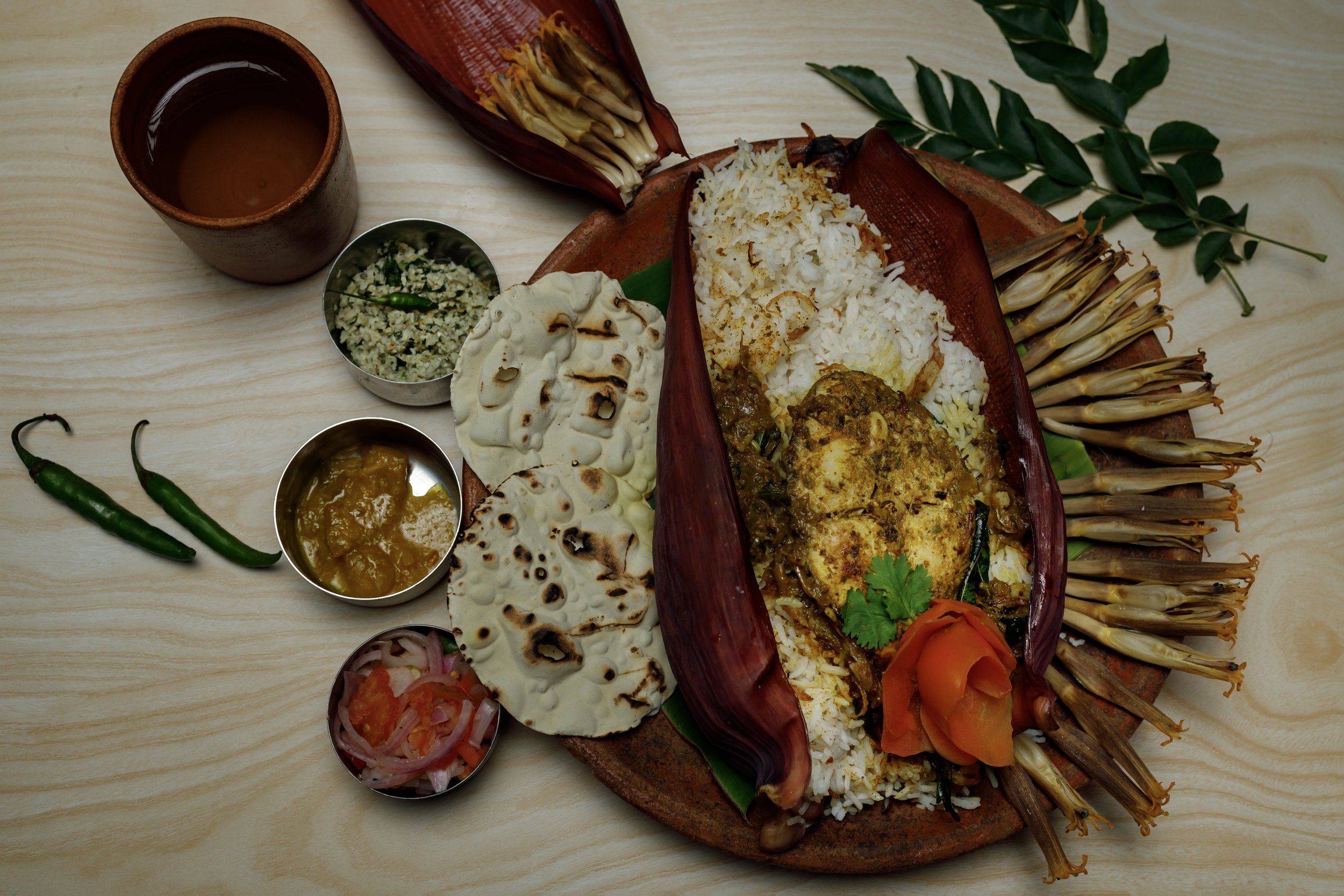 A traditional Indian thali with rice, curry, chapati, and side dishes, served on a wooden table with green chilies, a cup of tea, and garnishes.