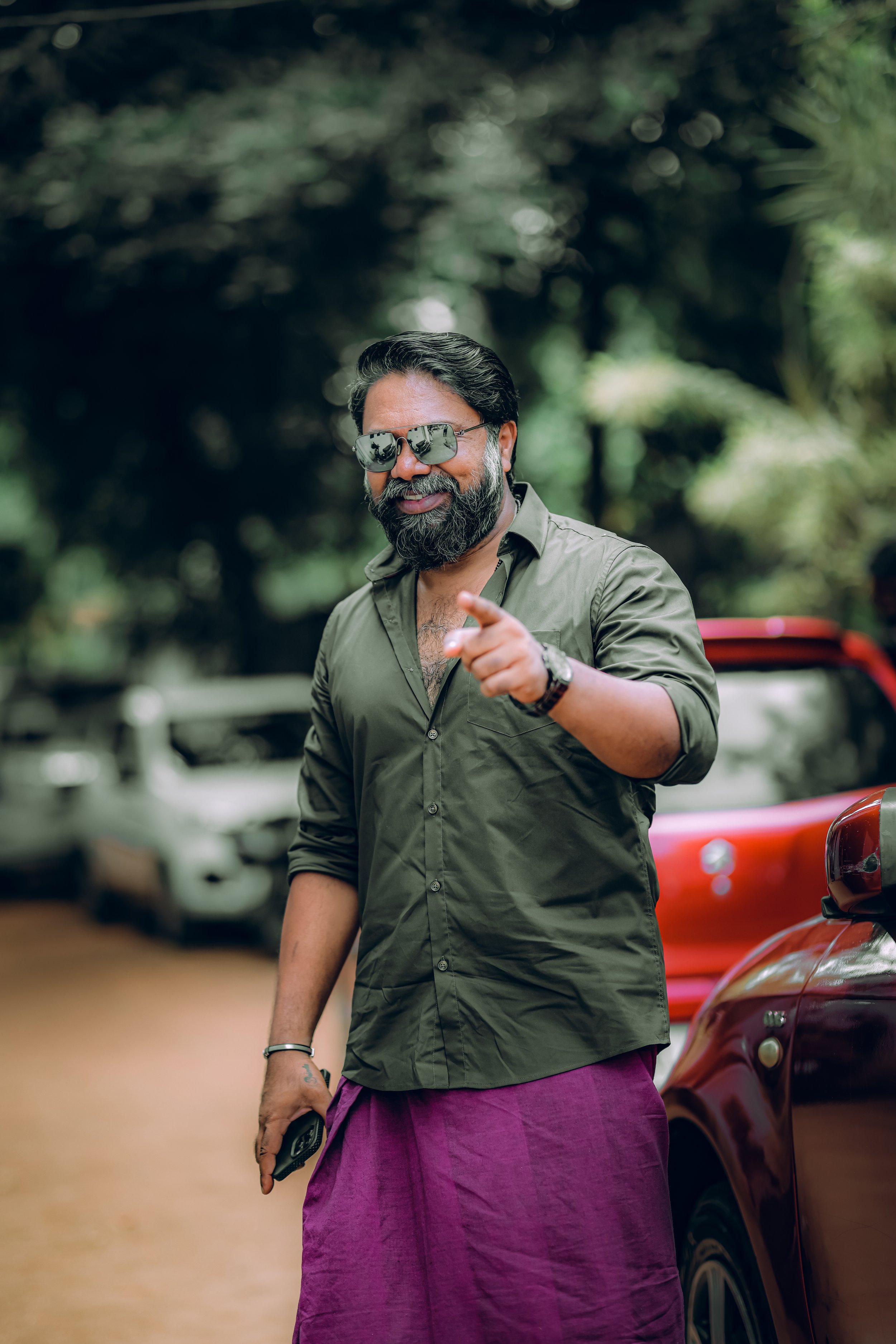 A bearded man wearing sunglasses, a green shirt, and a purple sarong, pointing at the camera while holding a phone, standing outdoors near cars with trees in the background.