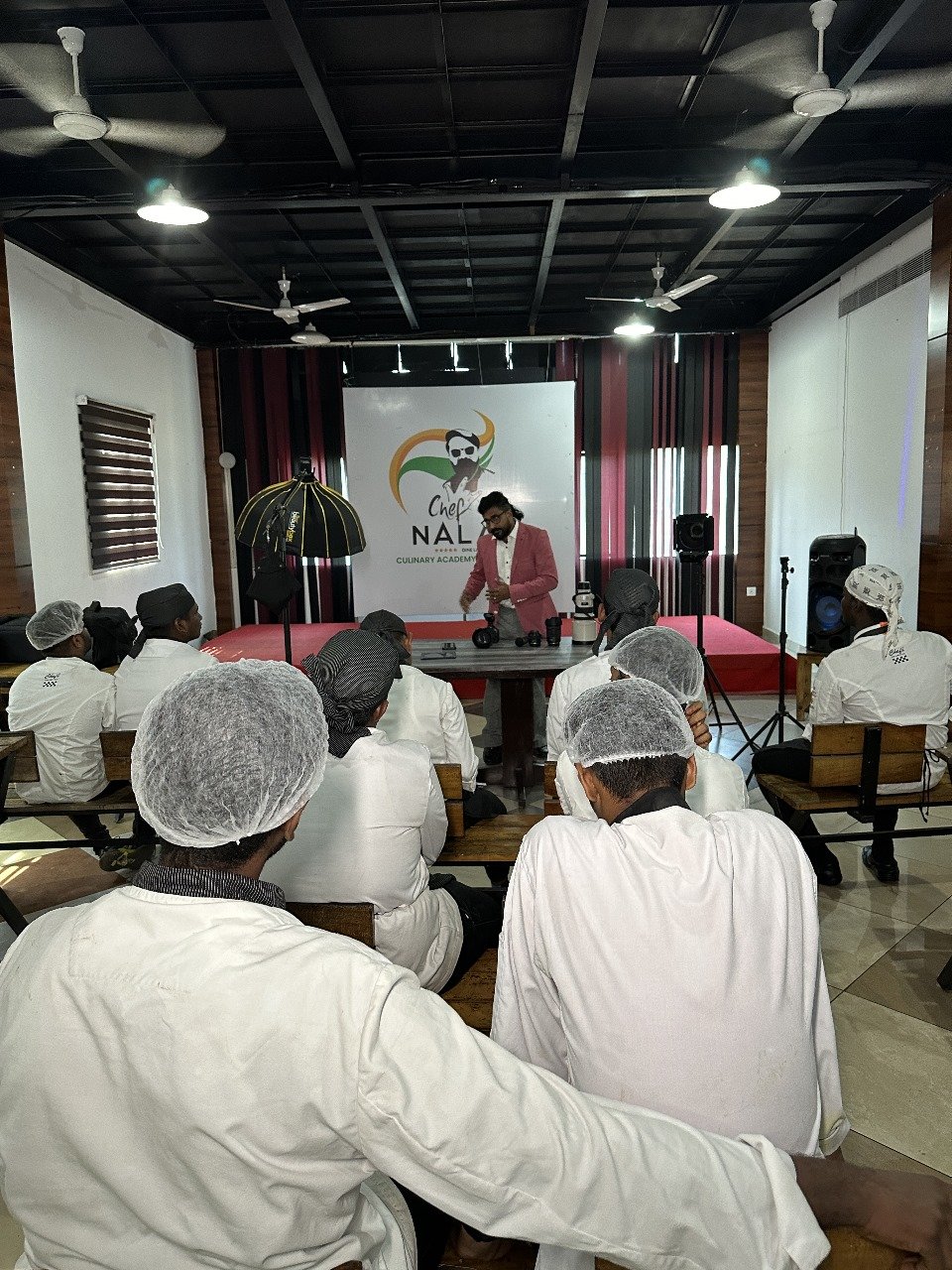 Cooking class with multiple students in white uniforms and hairnets, instructor in pink blazer demonstrating techniques on a table, in a room with a stage and chef logo backdrop.