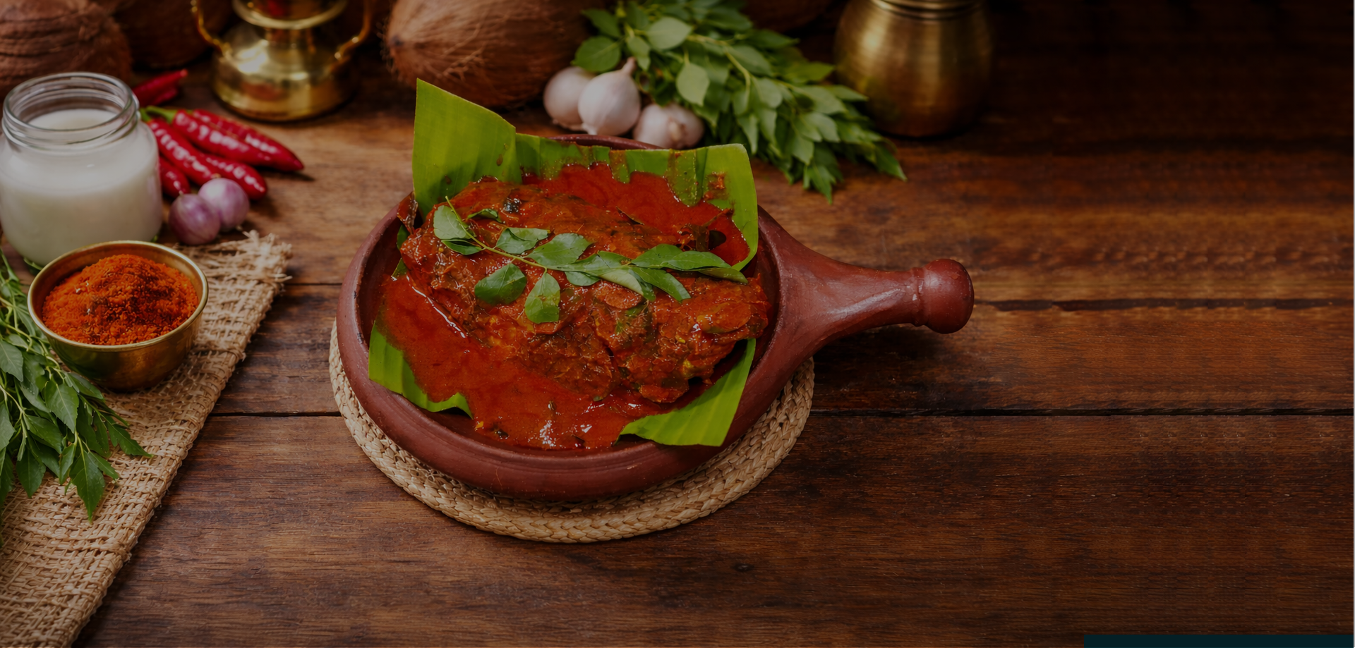 A traditional Indian dish served in a brown clay pot, topped with green curry leaves and covered with chili sauce, placed on a woven mat on a wooden table surrounded by ingredients like garlic, red chilies, spice powder, and greens.