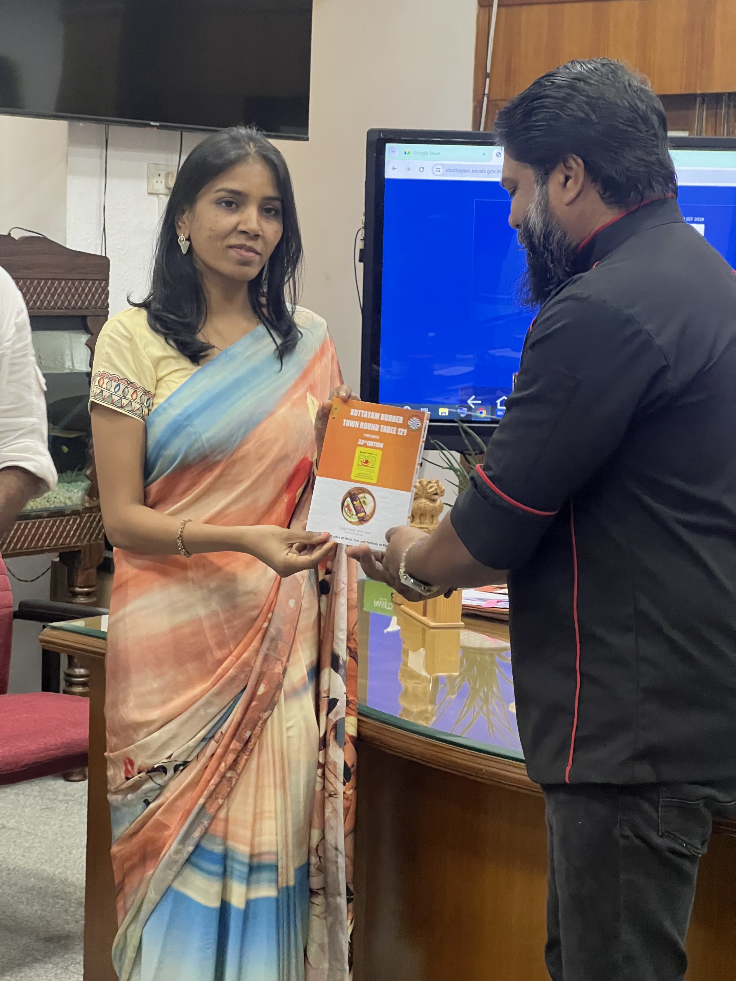 A woman in a patterned saree receives a booklet from a man in a black shirt during an indoor event, with a large monitor in the background.