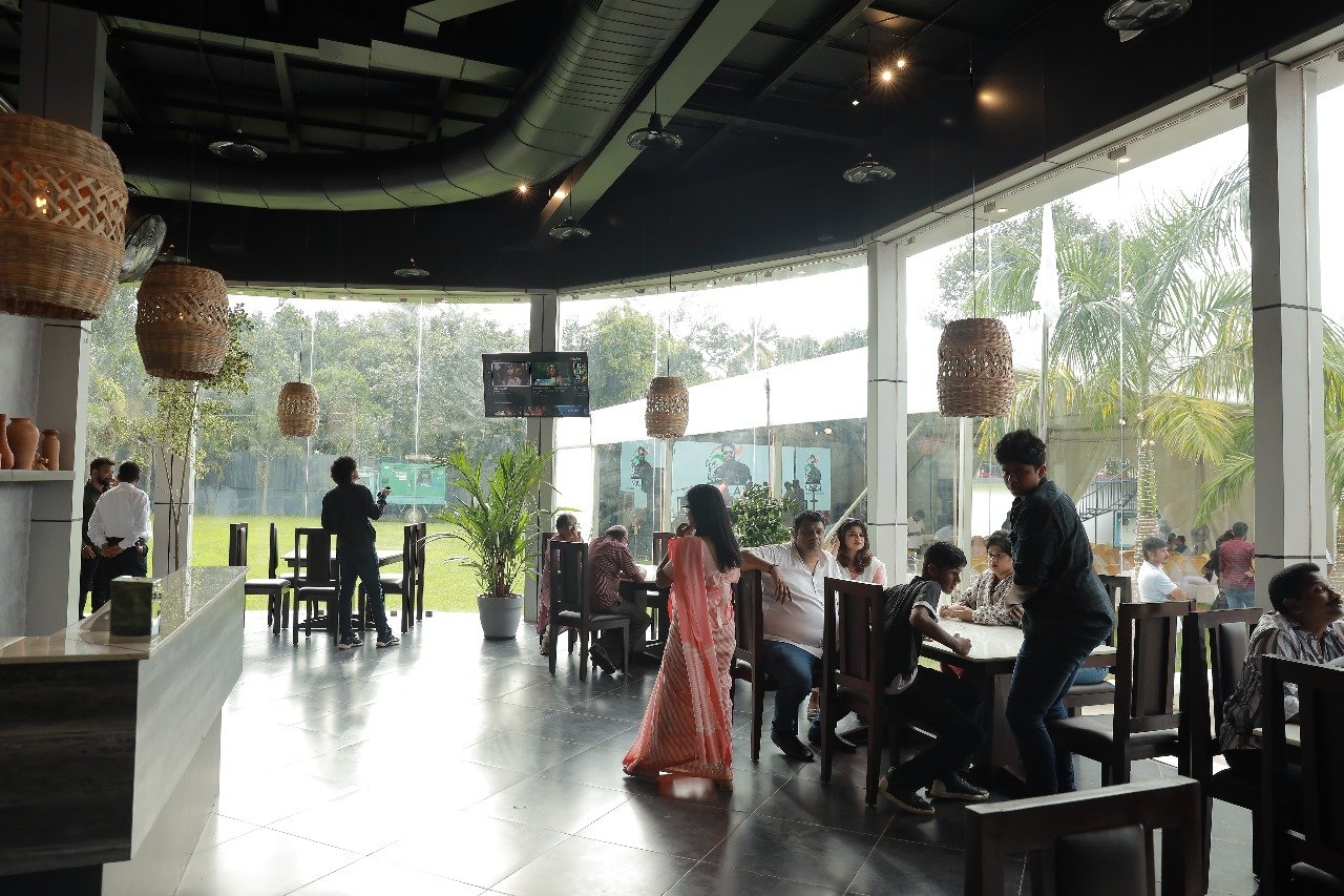 Indoor restaurant with large glass windows, wooden hanging lamps, and several people dining and socializing.