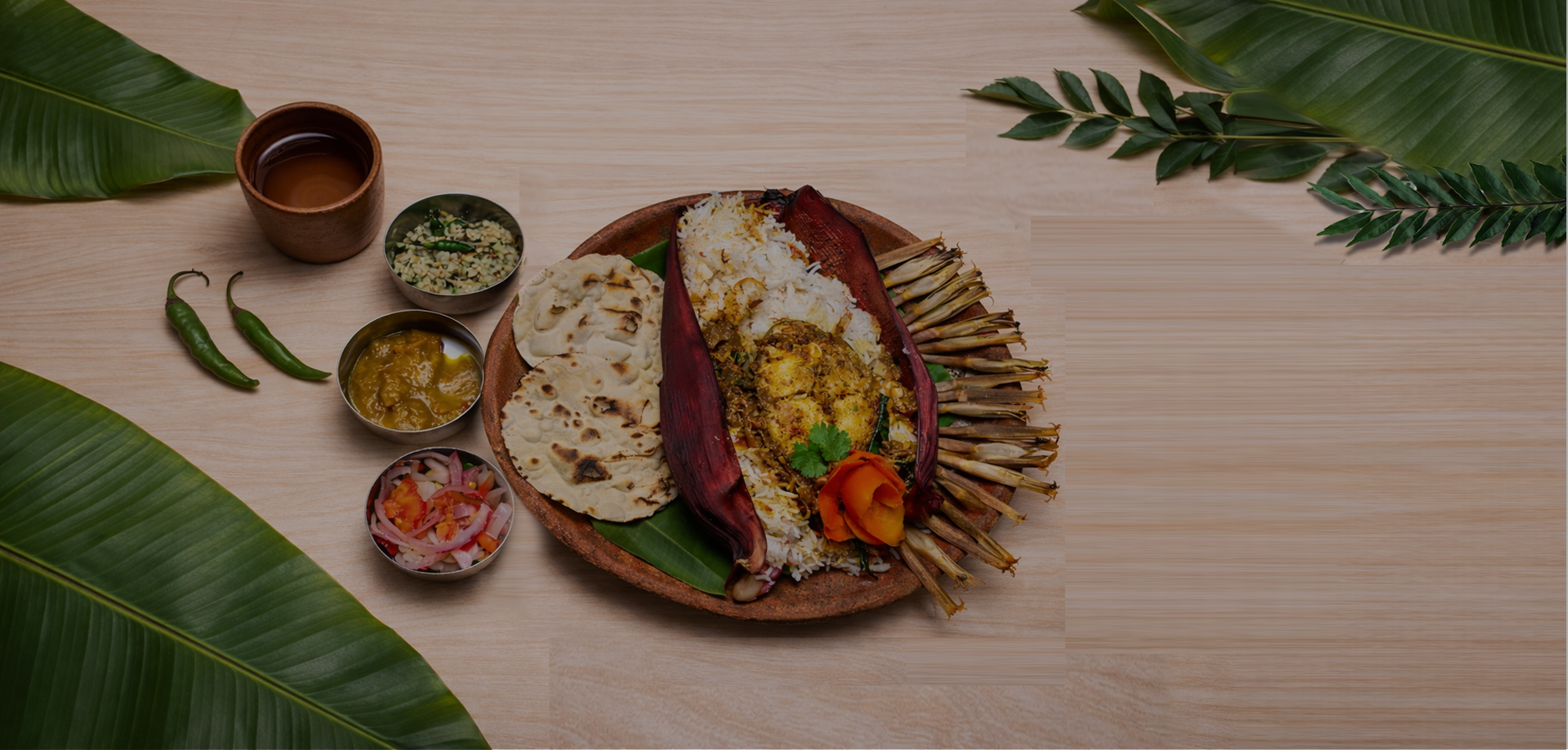 Plate of Indian food with rice, vegetables, chapati, and grilled prawns surrounded by side dishes and green leaves.