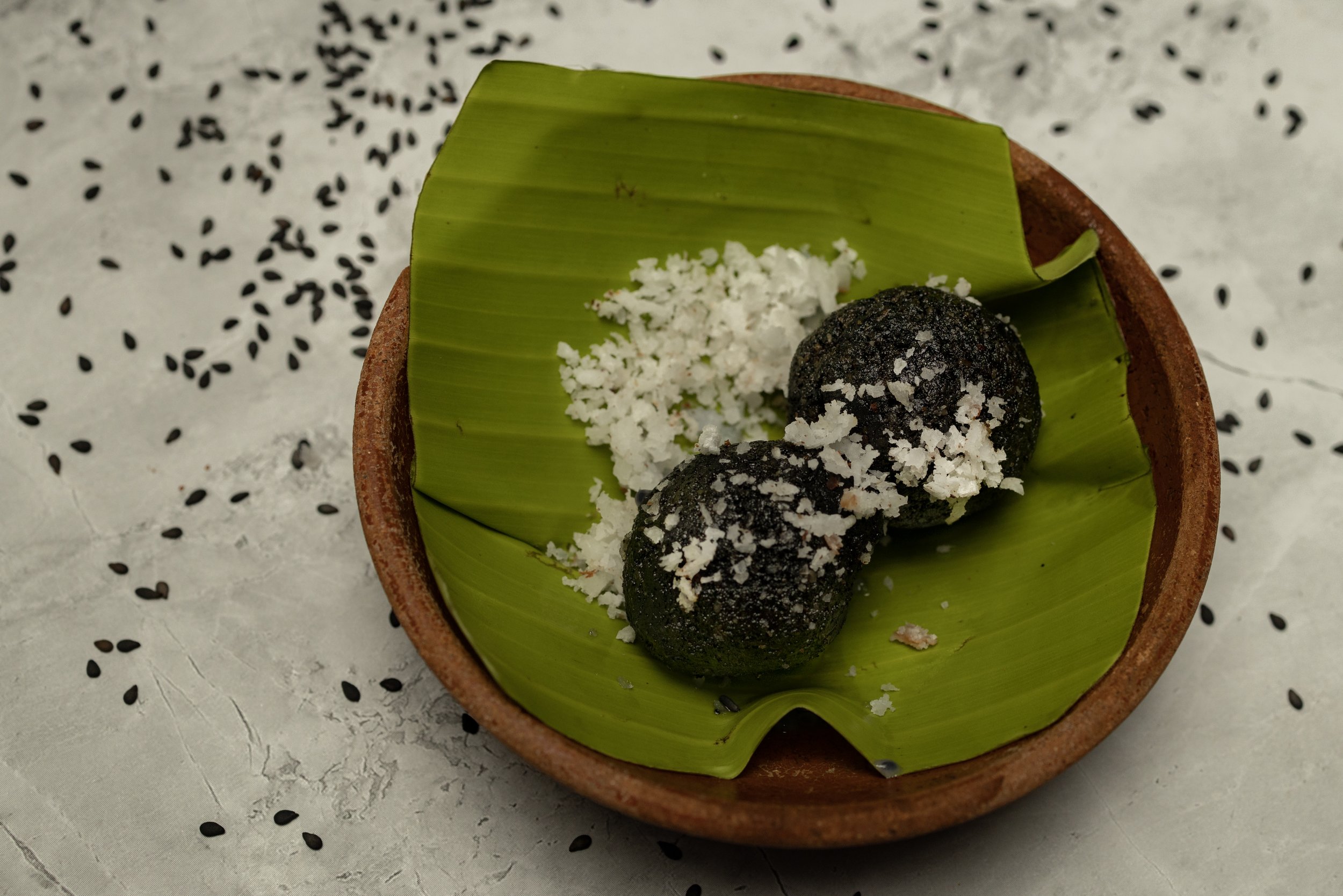 Two black round Indian sweets topped with grated coconut on a green leaf in a small brown bowl, with scattered black sesame seeds on white surface.