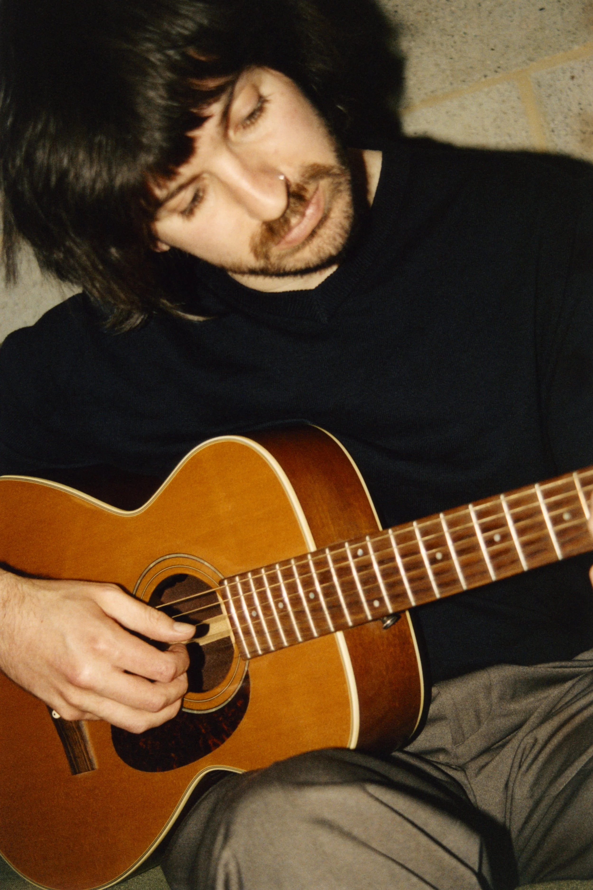 A man with dark hair, a beard, and a nose ring playing a wooden acoustic guitar, focusing on his hand and the guitar's fretboard.