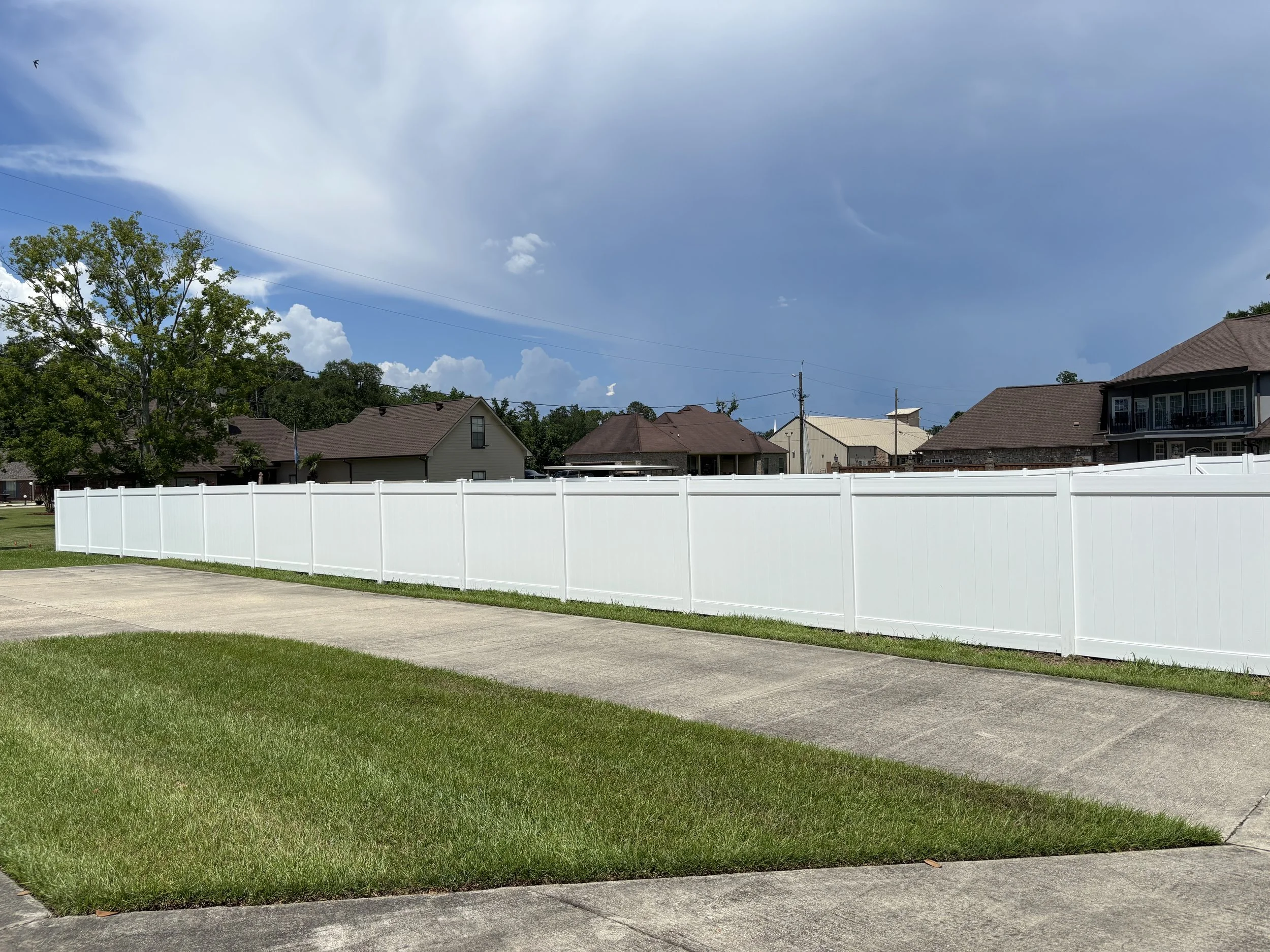 A white vinyl privacy fence along a sidewalk and grassy yard with residential houses and a large tree in the background, under a partly cloudy sky.