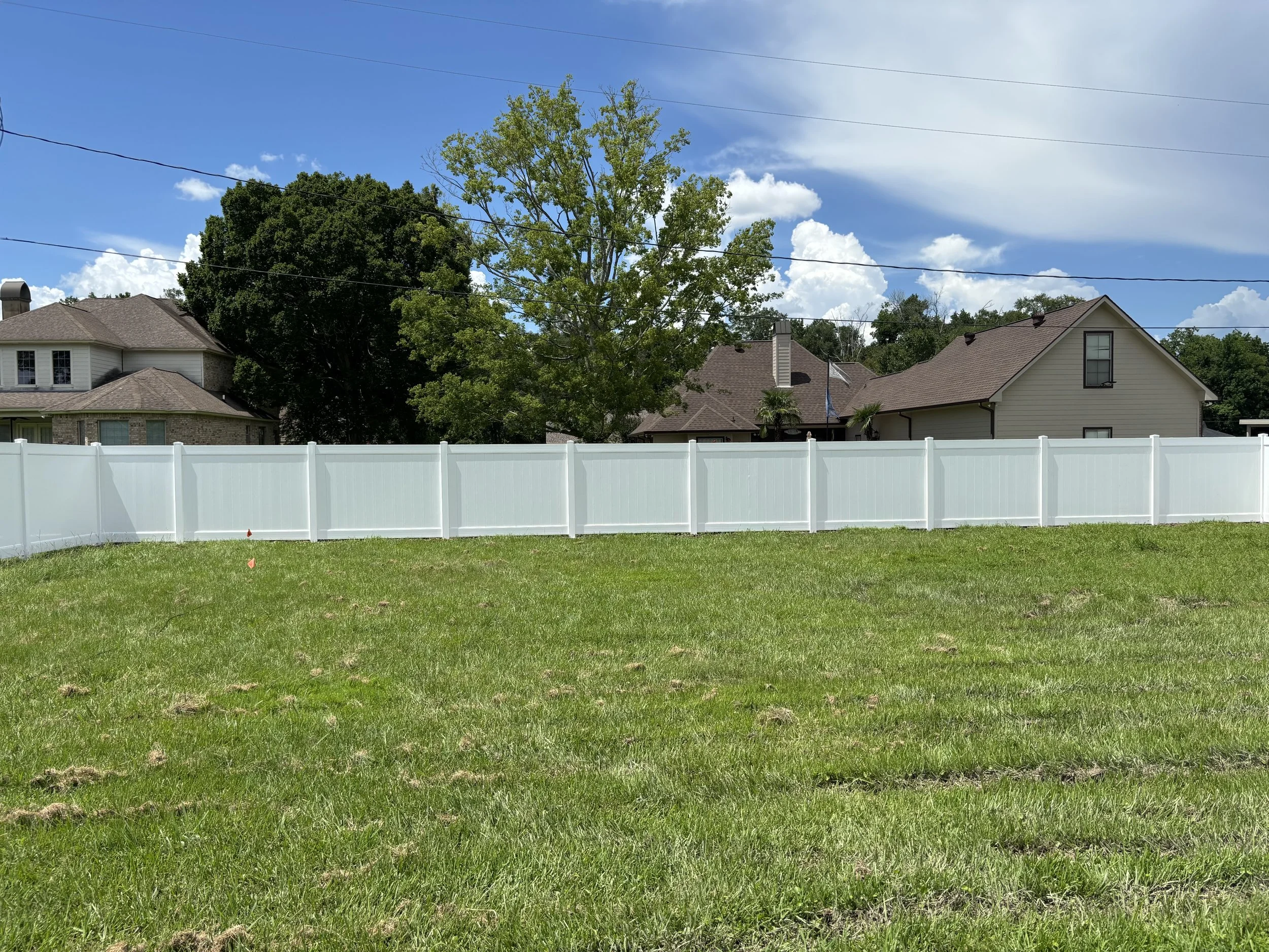 A backyard with green grass, a white vinyl fence, and neighboring houses with trees under a partly cloudy blue sky.