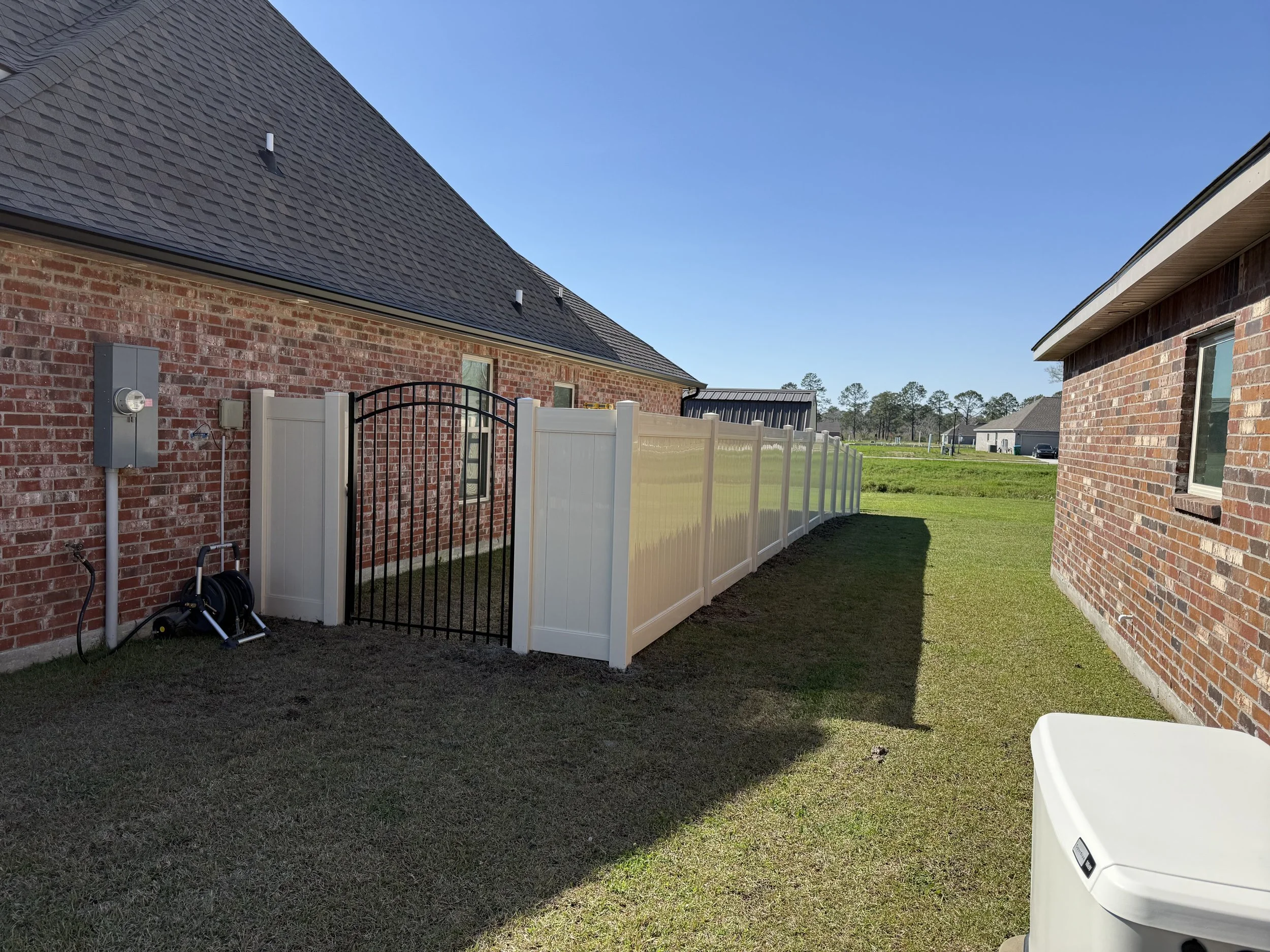 View of a backyard with a white vinyl fence, a brick house on the left, a brick house on the right, and a grassy open field in the background under a clear blue sky.