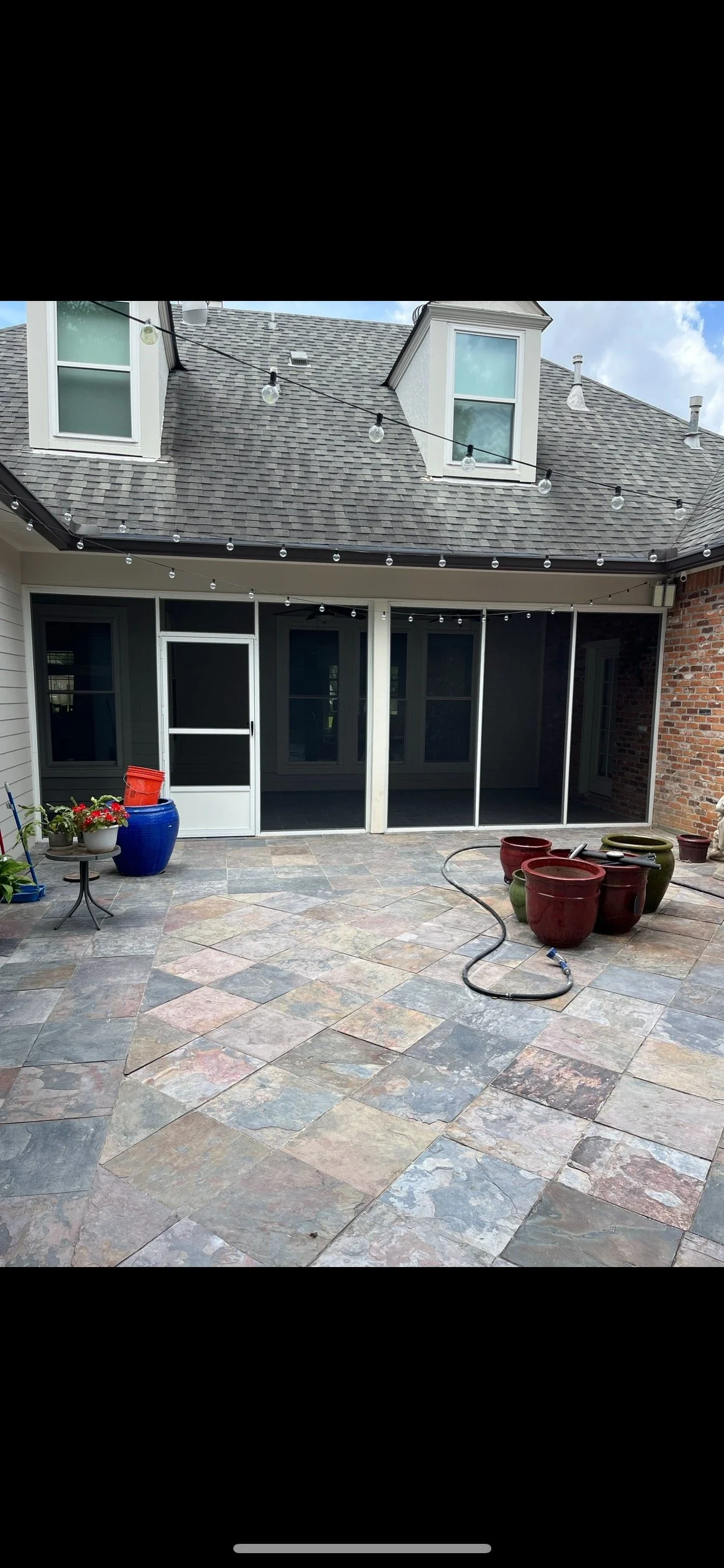A screened-in patio area with stone tile flooring, potted plants, and string lights above, attached to a house with gray siding and a brick wall.