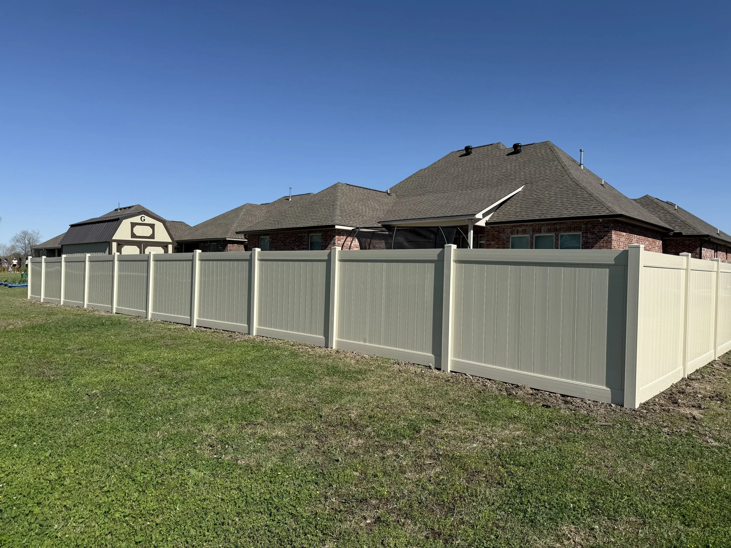 A backyard with a beige vinyl privacy fence enclosing a brick house with a grey shingled roof. The sky is clear and blue.