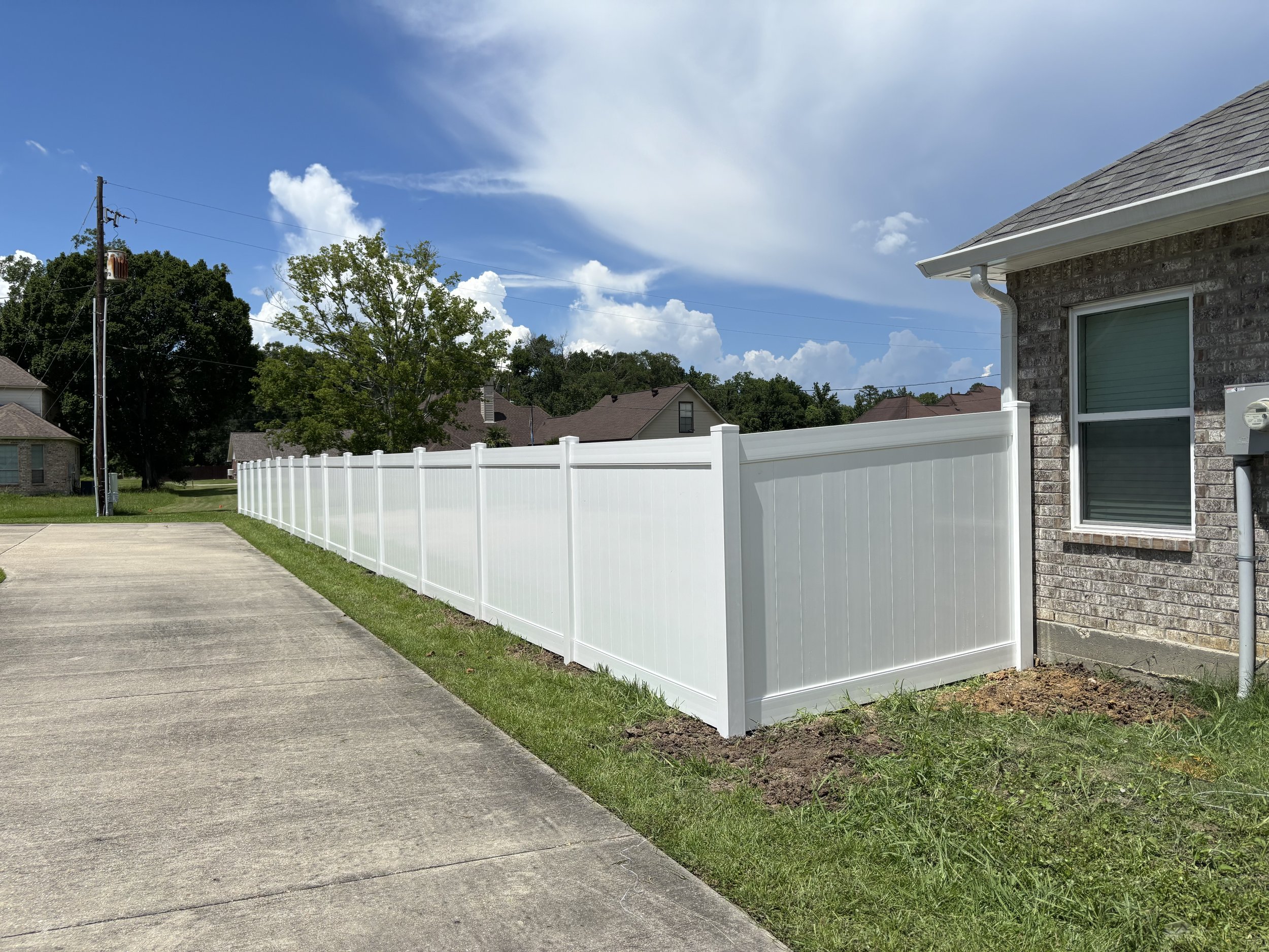 A white vinyl fence runs alongside a brick house with a gray roof, adjacent to a concrete driveway and a small patch of grass. The sky is partly cloudy with some trees visible in the background.