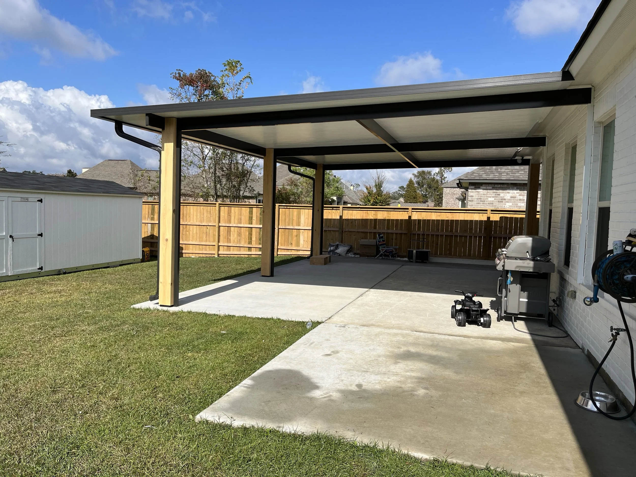 A backyard patio with a newly built covered area, concrete flooring, a white brick house on the right, a wooden fence in the background, and a garden shed on the left. There is a grill, some tools, and a small toy vehicle on the patio.