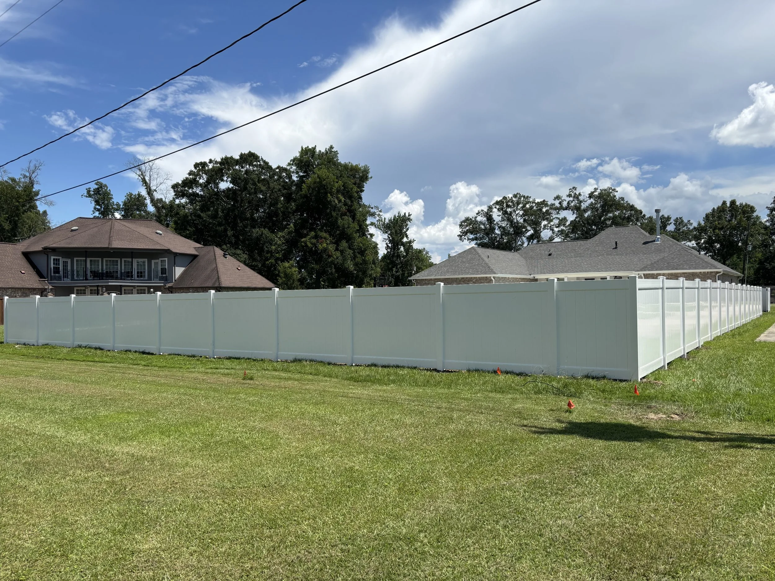 Residential backyard with a white privacy fence, two houses with gray and brown roofs, green grass, leafless trees in the background, and a partly cloudy sky.
