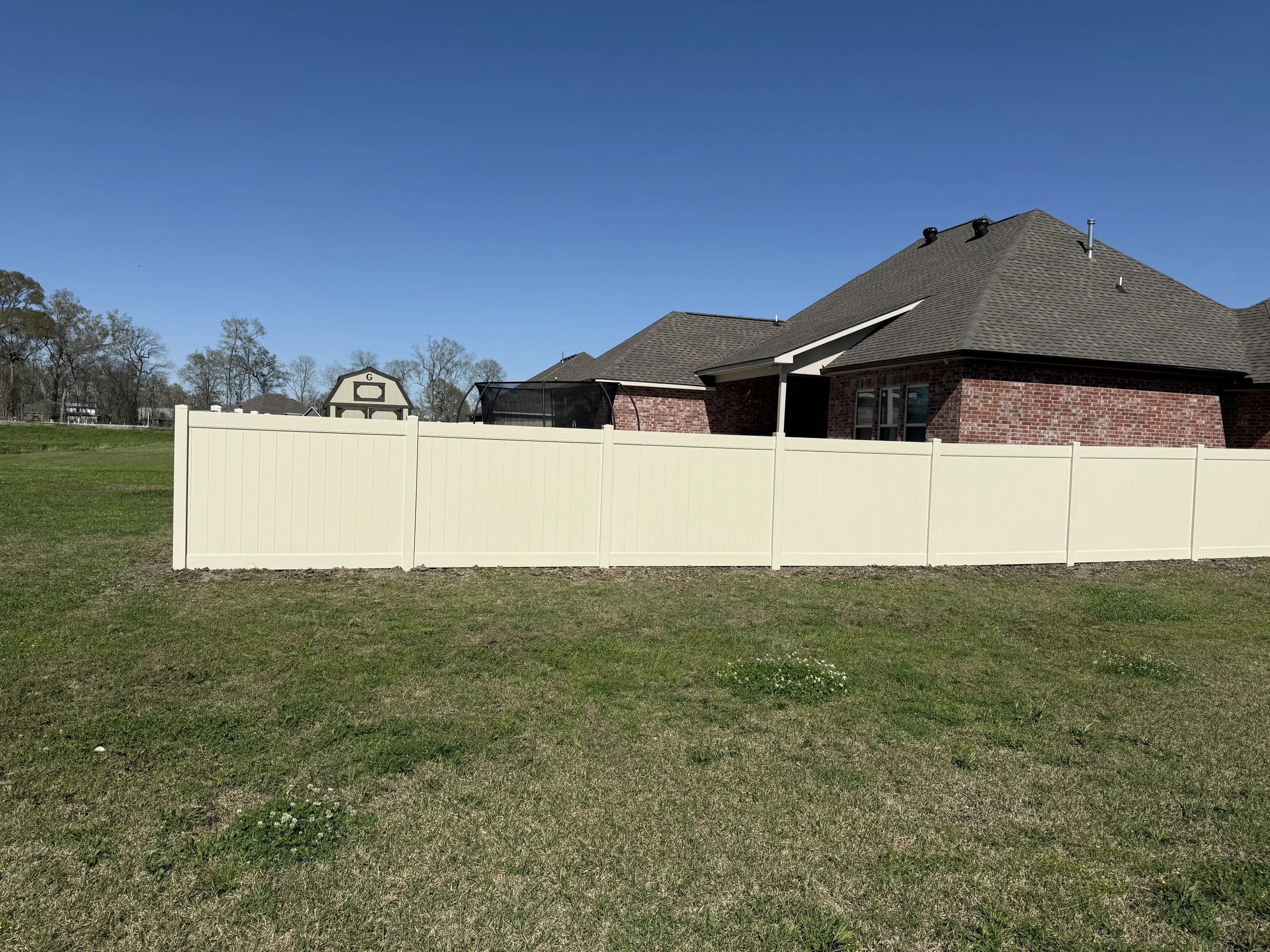 Residential backyard with a white vinyl fence, brick house with a gray shingle roof, and a lawn with patches of grass. In the background, there are trees and a clear blue sky.