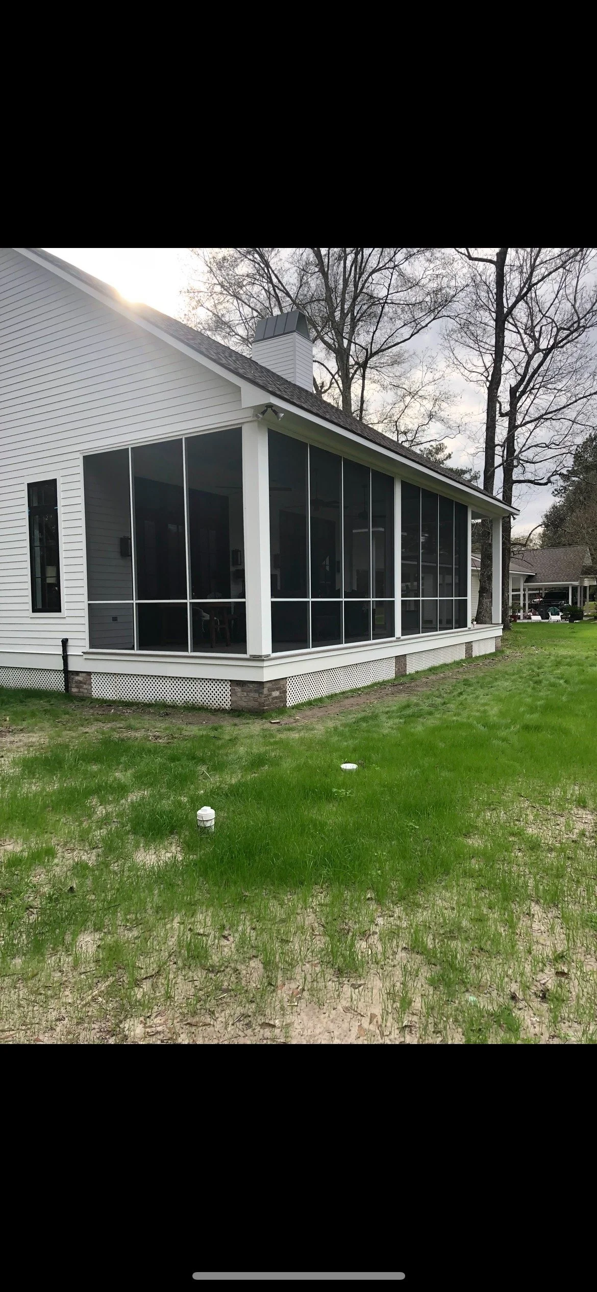 A house with a screened-in porch, white siding, and brick foundation, with a grassy yard and trees in the background.