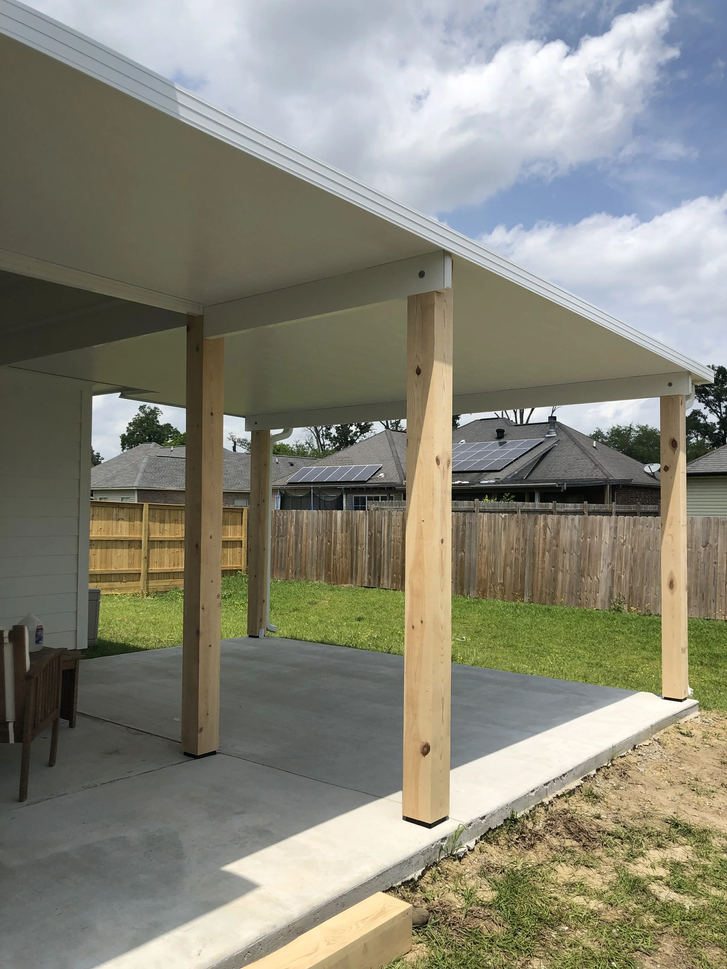 Construction of a new porch or patio with wooden posts and a concrete slab in a backyard, with neighboring houses and solar panels visible in the background.