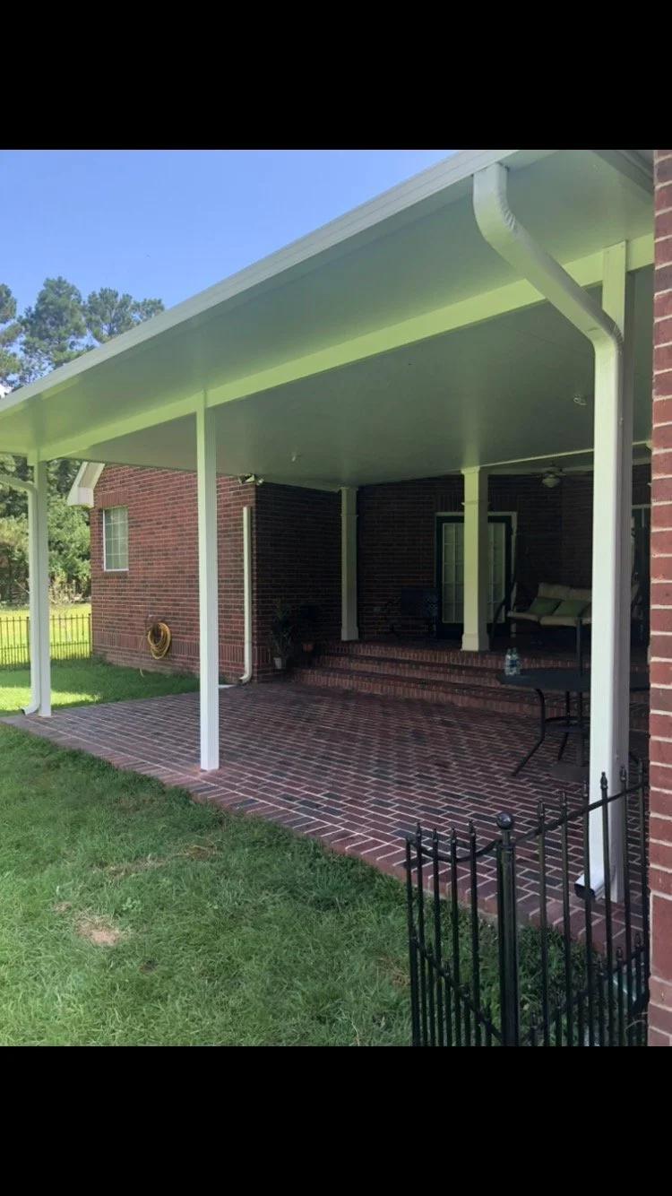 Back patio area with brick flooring, covered by a white roof supported by white columns, attached to a brick house. There is outdoor furniture, including a couch, and a small table with a water bottle. A grassy yard surrounds the patio, and a garden 