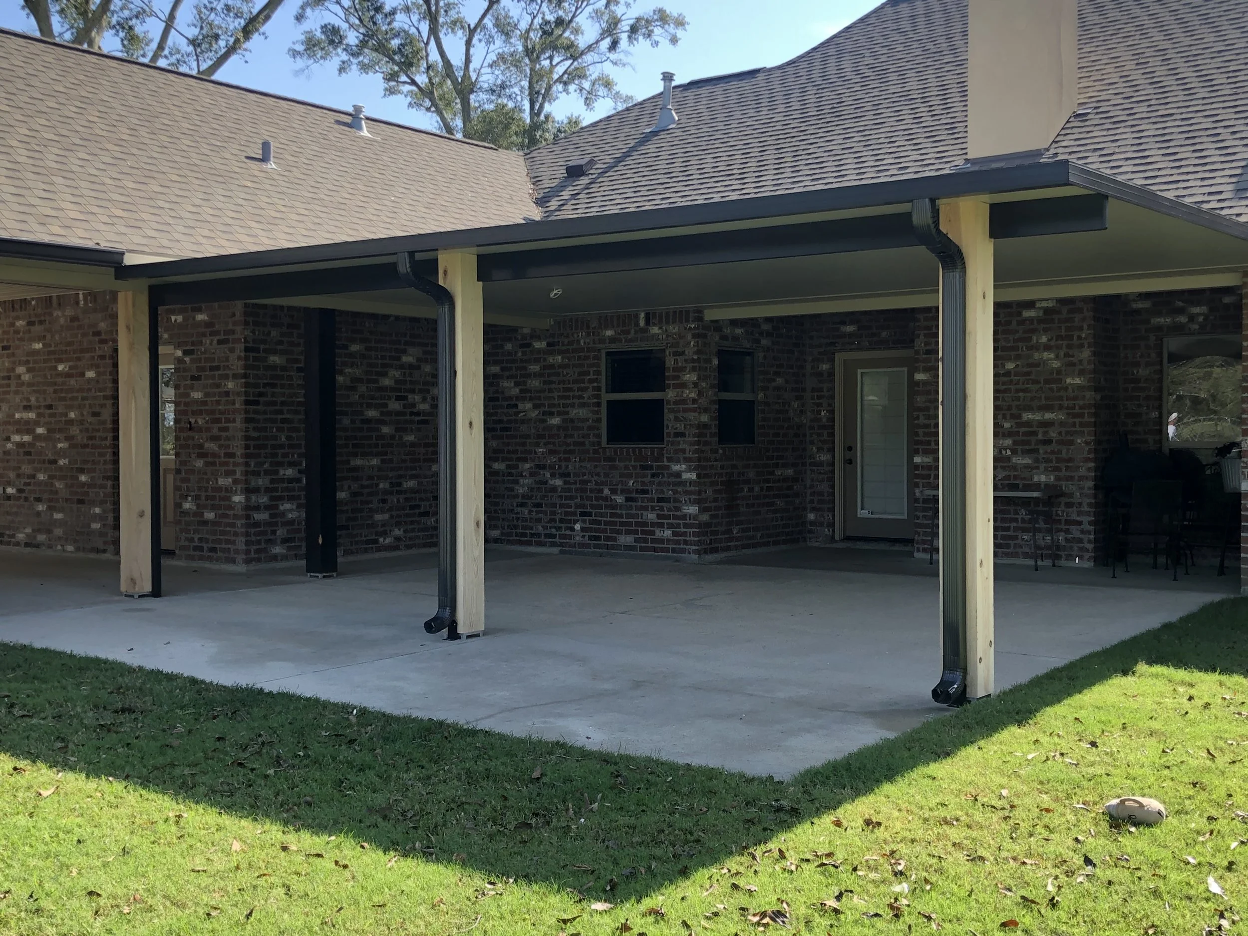 Backyard patio with brick house, concrete floor, and a screened in porch area. Green grass surrounds the patio, and there is a black outdoor furniture set in the corner.