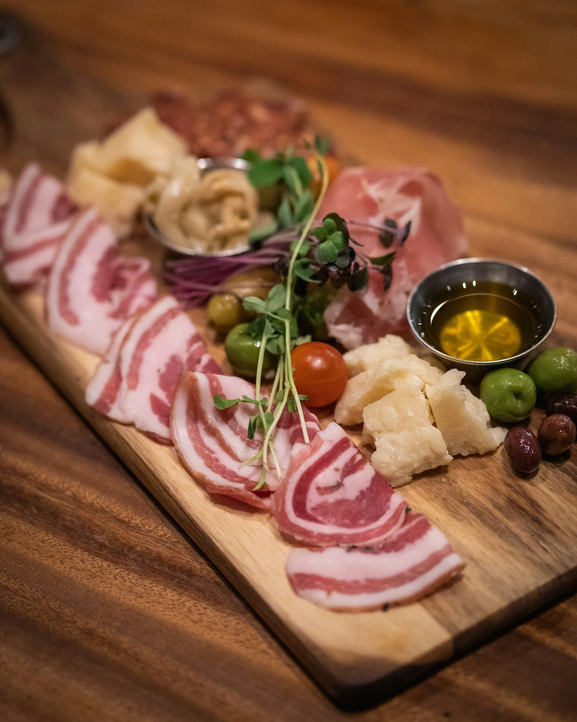 Plate de charcuterie avec différentes viandes, fromages, olives, tomates cerises, microgreens et huile d'olive sur une planche en bois.