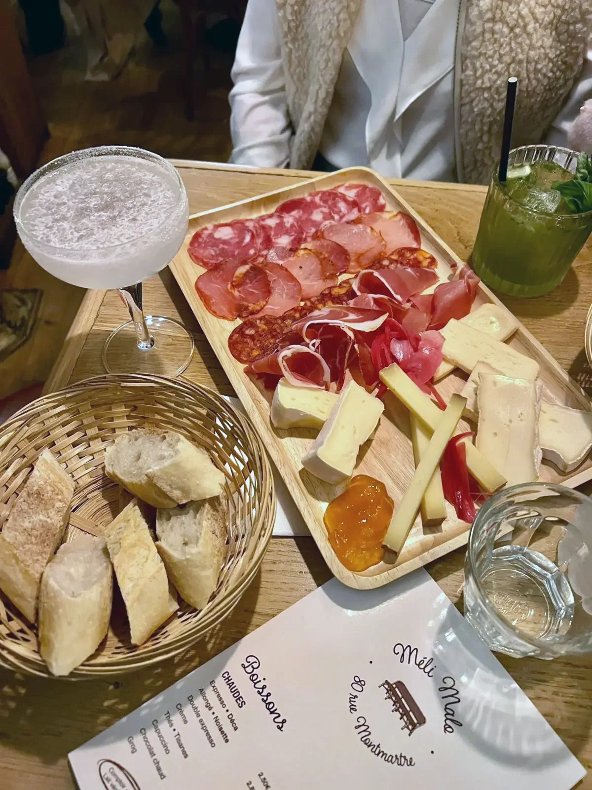 Un plateau avec une sélection de charcuterie, divers fromages, et un morceau de confiture. À côté, un panier avec du pain. Sur la table, un verre à cocktail avec une boisson claire, un verre de cocktail vert avec une paille et des feuilles de menthe, et une carafe d'eau. En arrière-plan, une personne en vêtement blanc et beige assise à une table en bois.