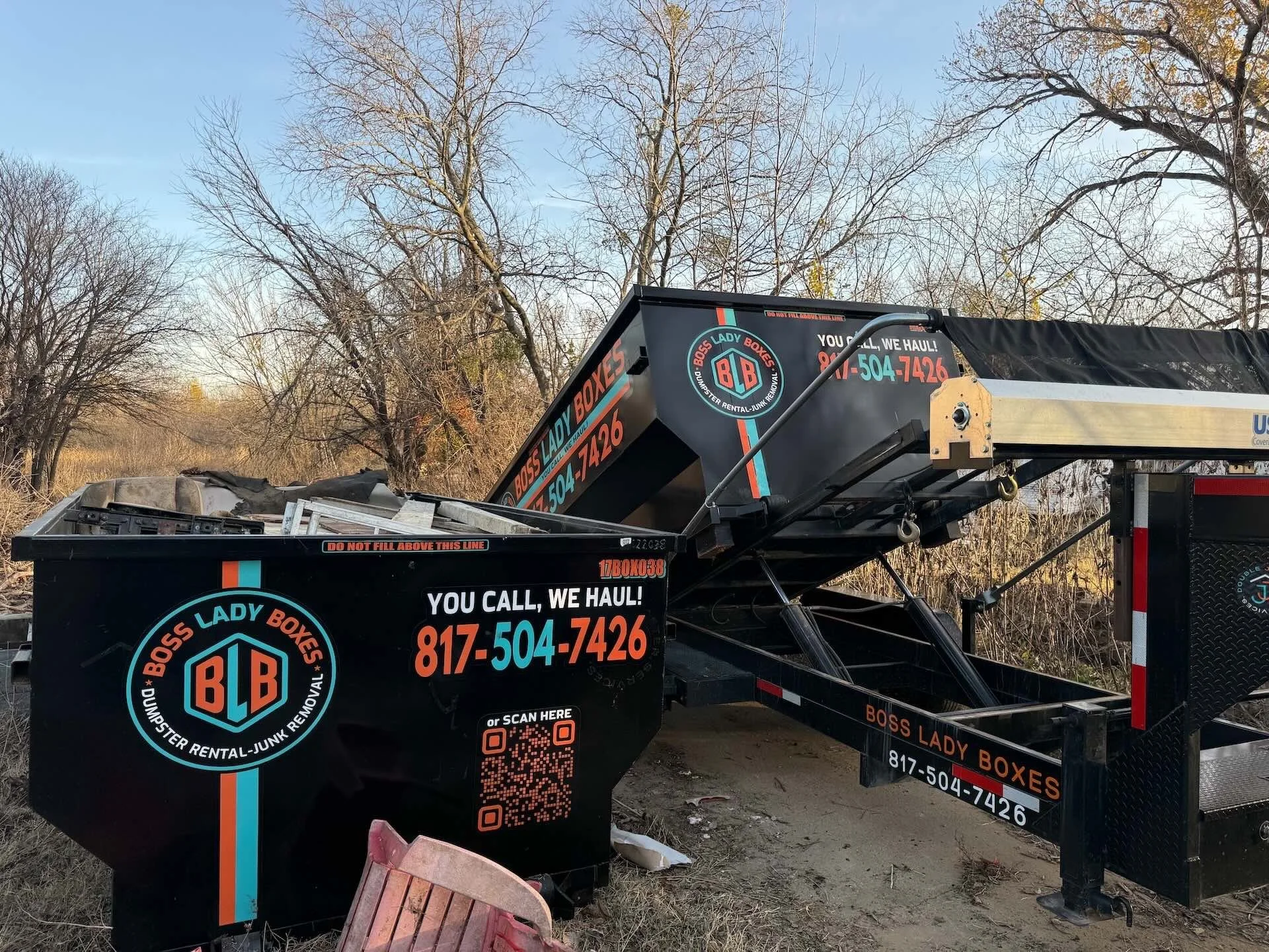 A black trailer with the logo and contact information of Boss Lady Boxes, a junk removal company, parked outdoors near leafless trees and bushes.